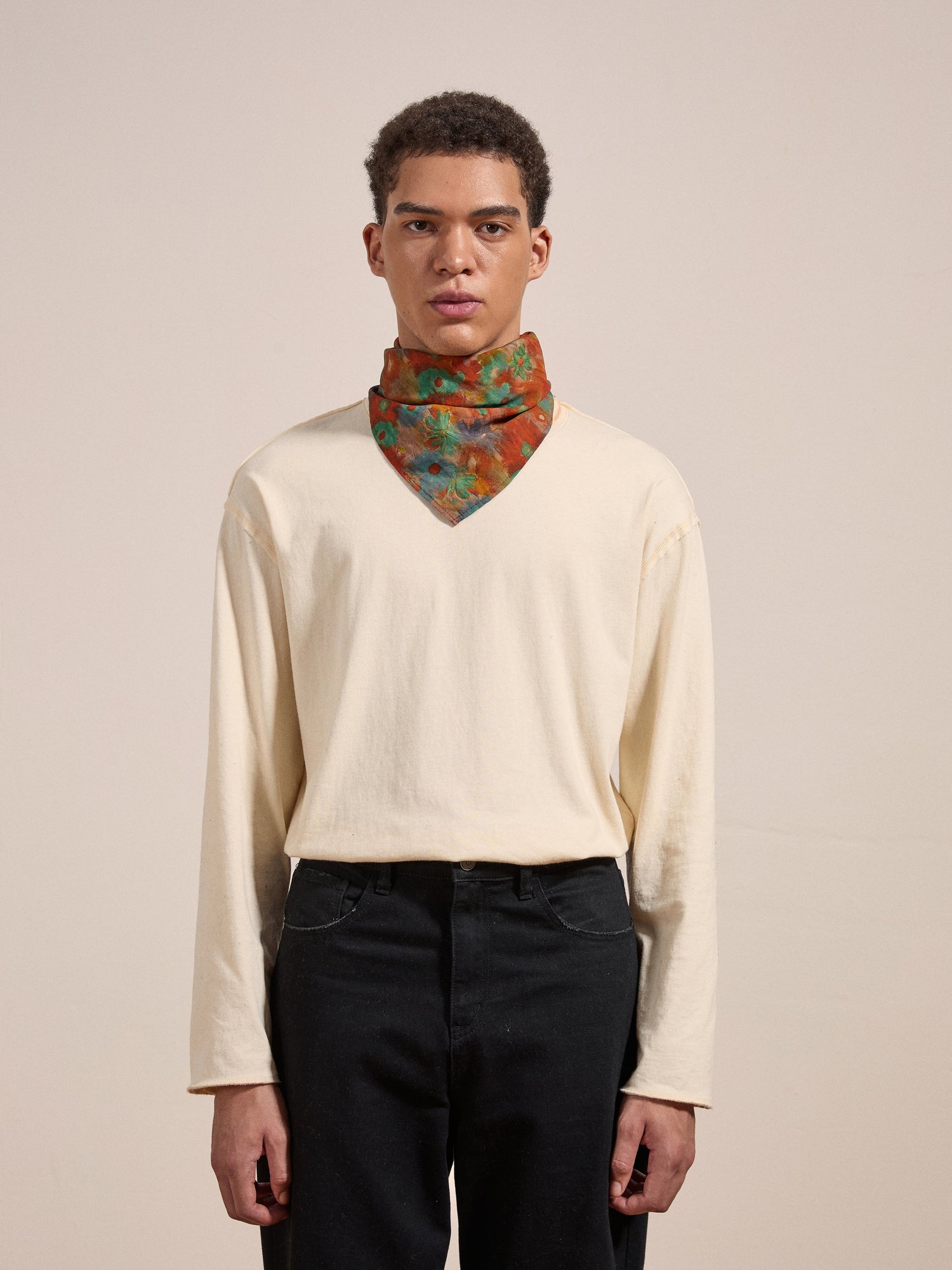 A person with short curly hair is wearing a cream-colored long-sleeve shirt, black pants, and the Sepehr Bandana from FOUND. They are standing against a plain, neutral background.