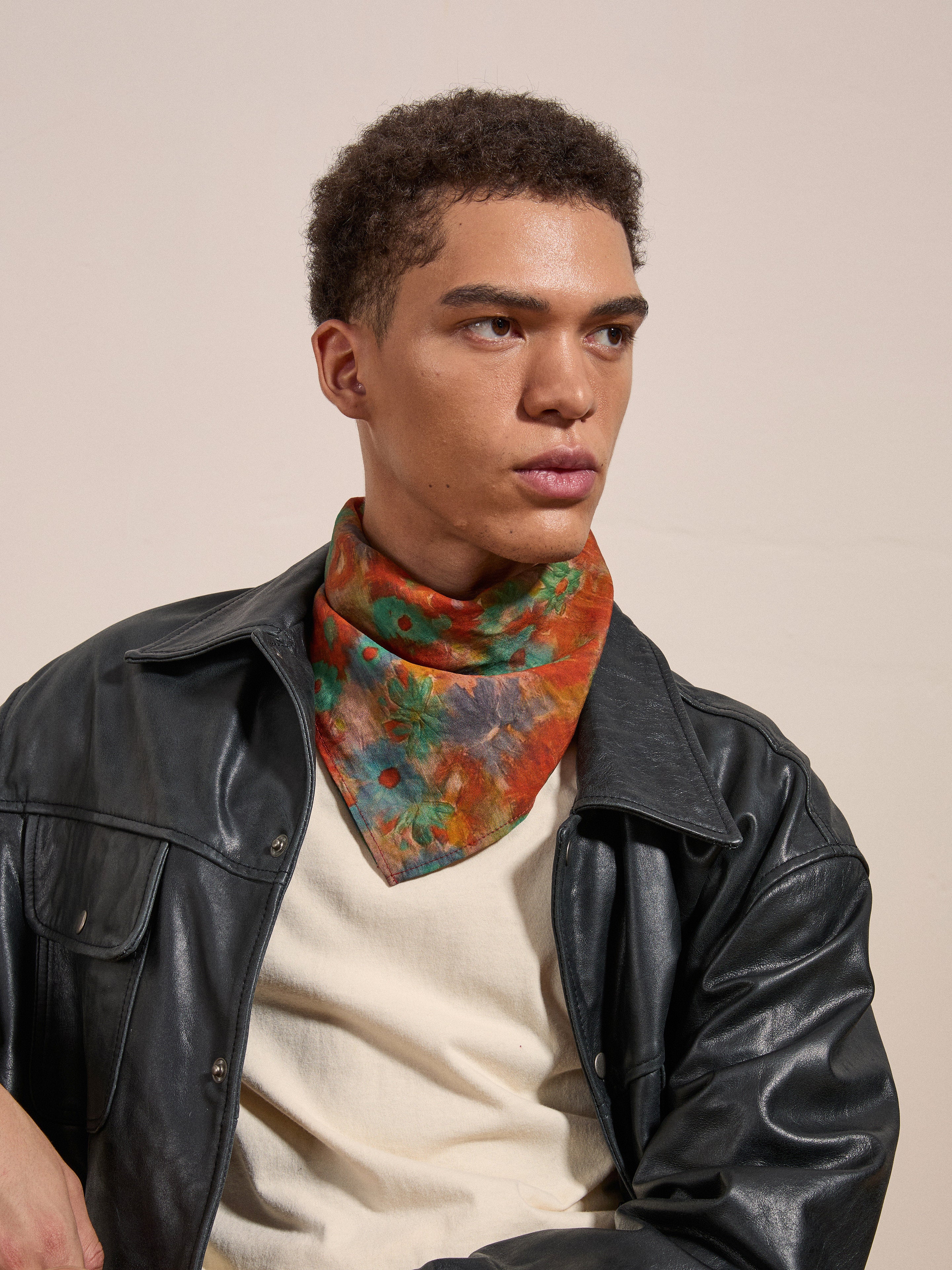 A person with short, curly hair wearing a black leather jacket, white shirt, and the Sepehr Bandana from FOUND, seated against a plain background.