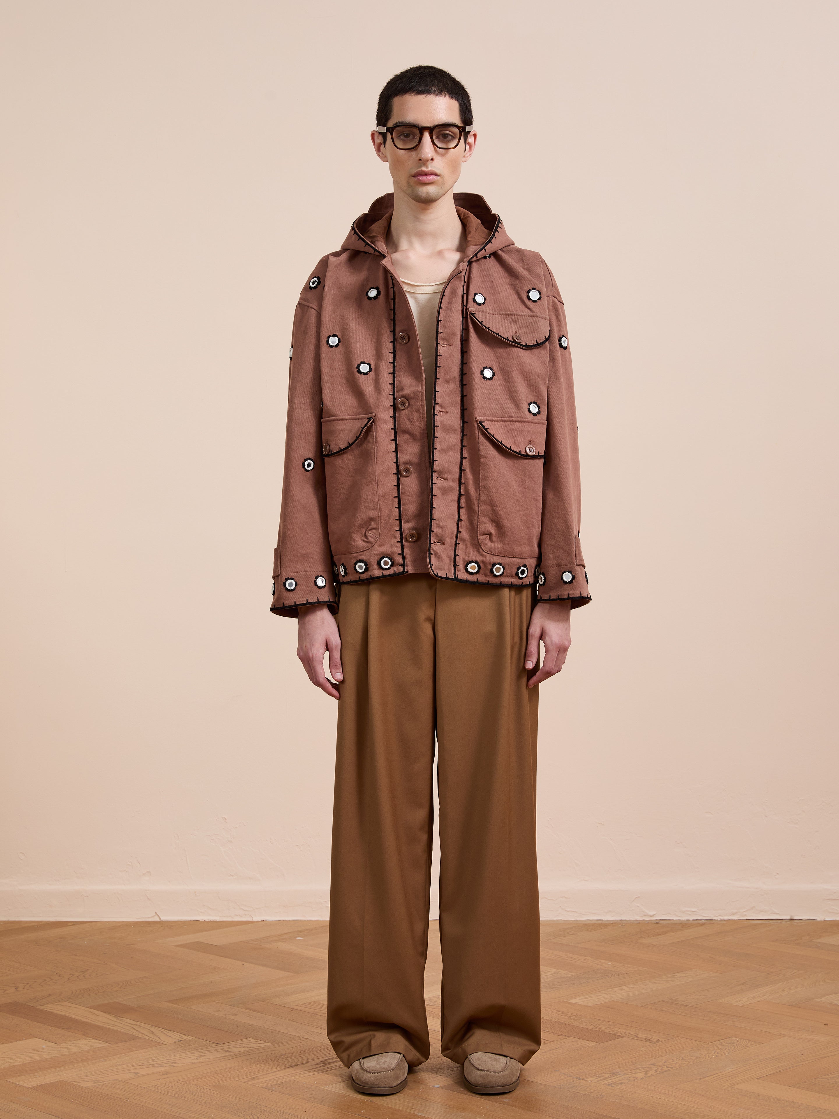 Someone stands indoors in a FOUND Hand Embroidered Hooded Mirror Anorak with large pockets and grommet details, matching wide-leg pants, beige shoes, and glasses against a plain light background.