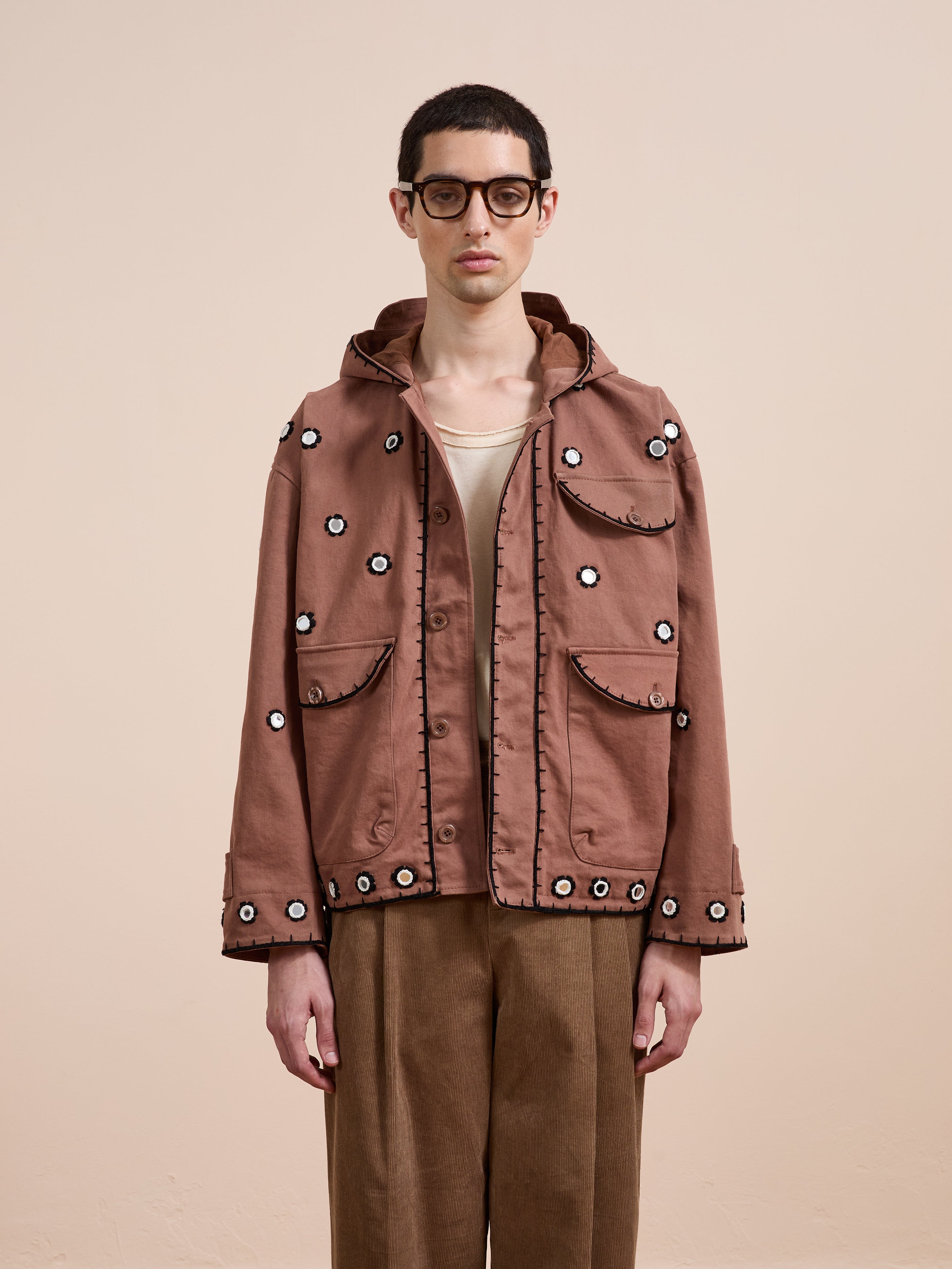 A person with short dark hair and glasses wears the FOUND Hand Embroidered Hooded Mirror Anorak with multiple pockets, paired with brown pants, against a plain beige background.