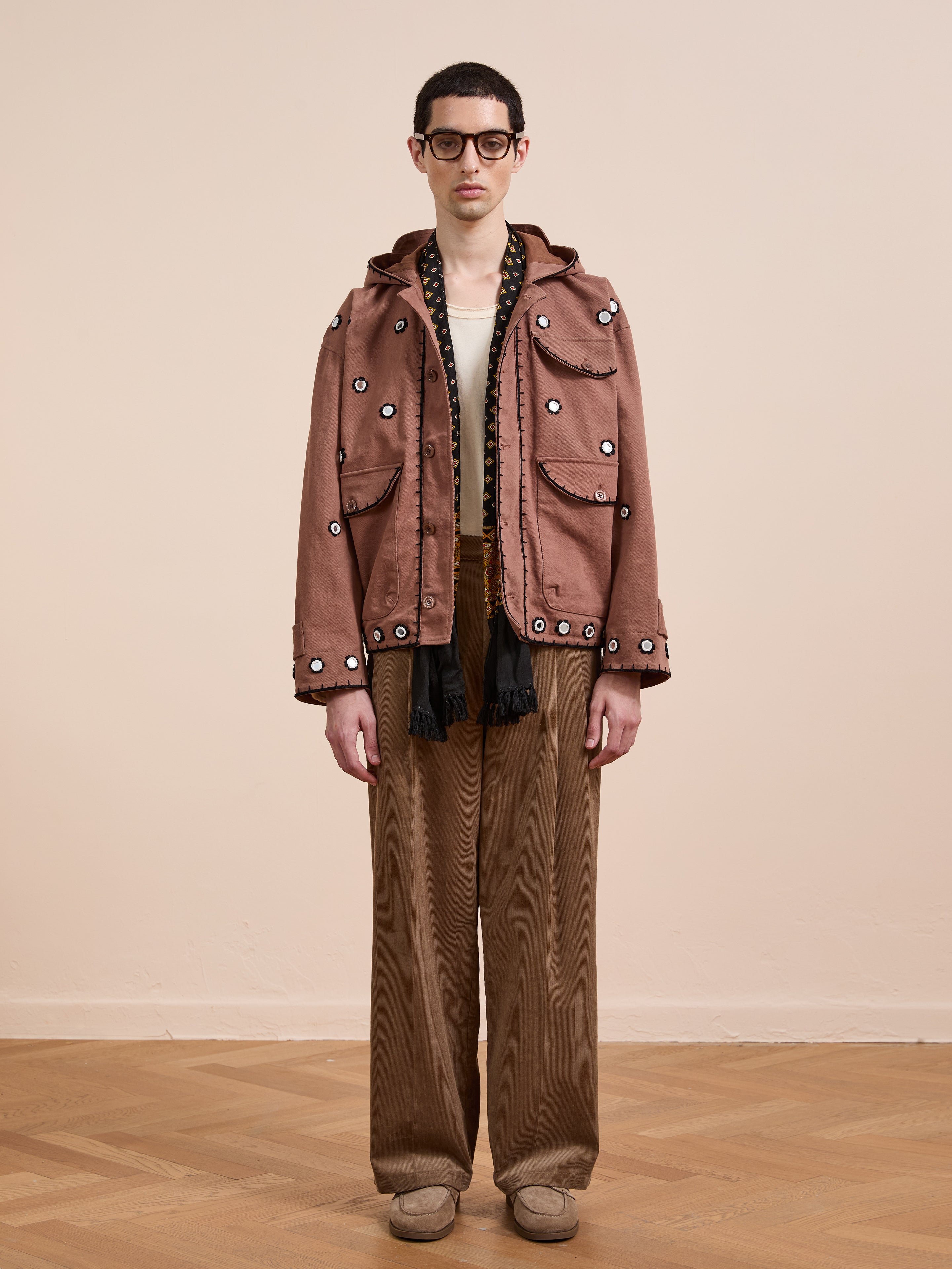 Indoors, a person wears FOUND’s Hand Embroidered Hooded Mirror Anorak, paired with glasses, a white shirt, brown wide-leg pants, and beige shoes against a plain background.