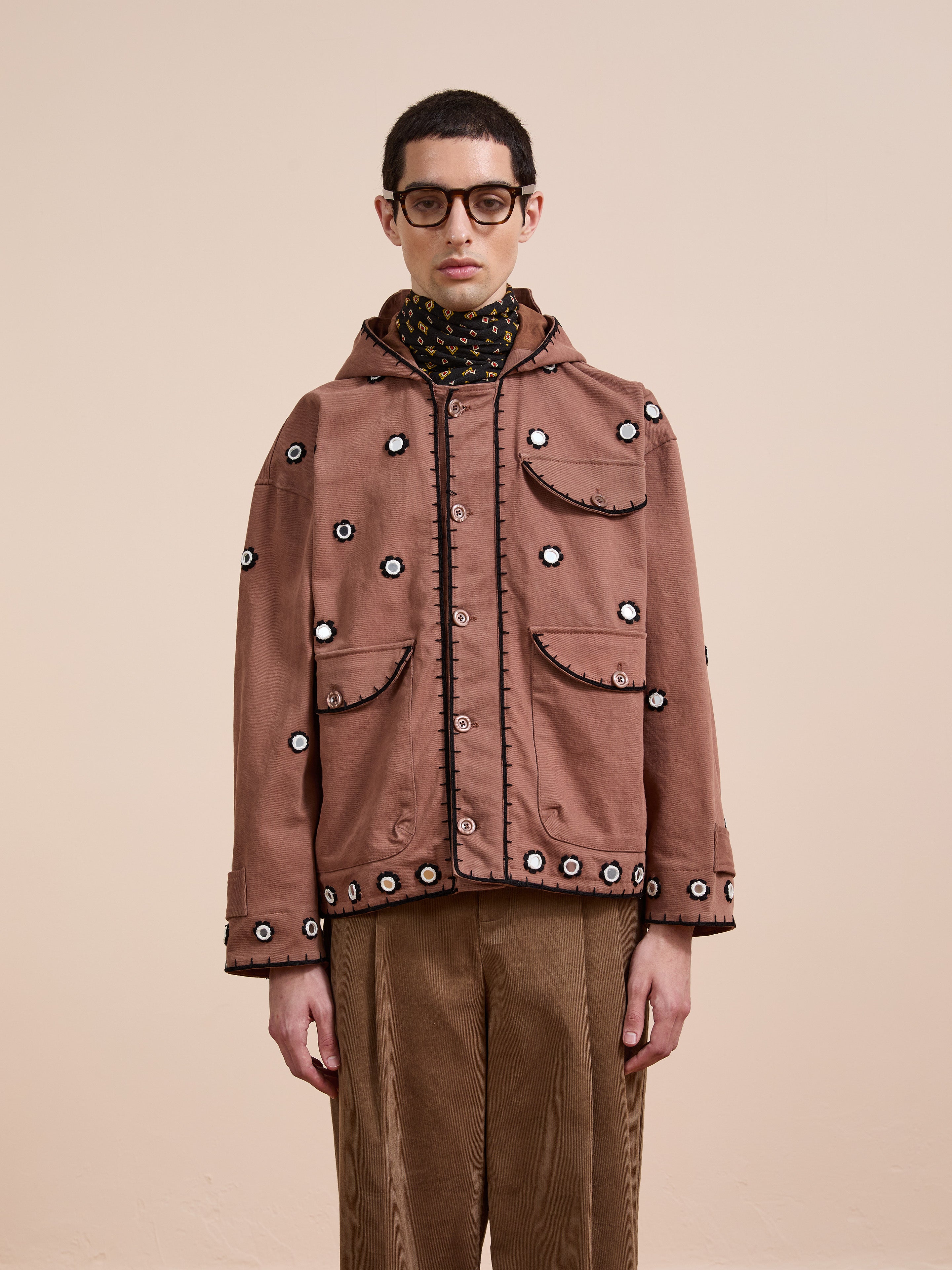 A person in FOUND's Hand Embroidered Hooded Mirror Anorak, glasses, a patterned turtleneck, and tan corduroy pants stands against a plain beige background.