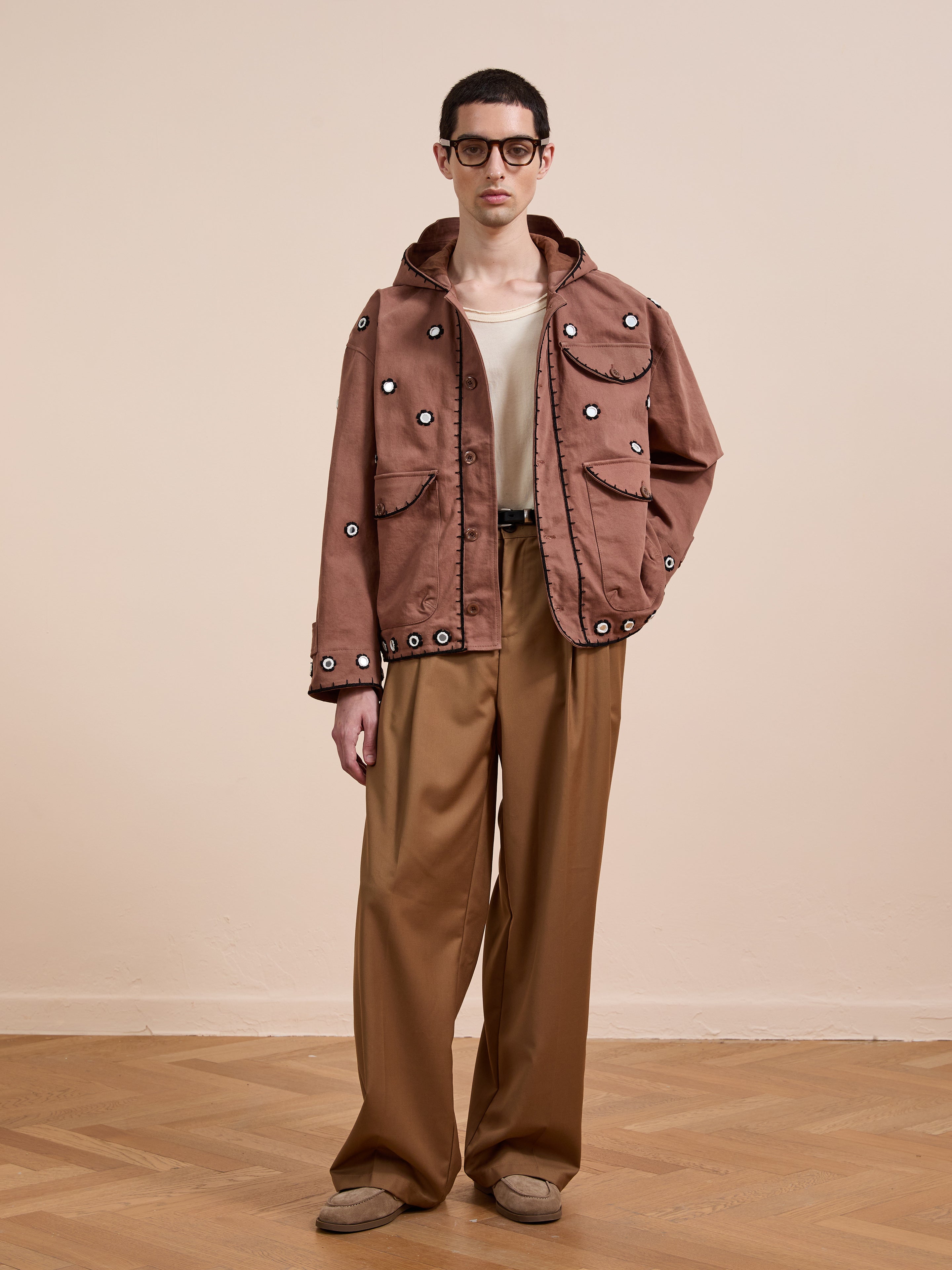 Indoors, a person wears glasses, a white shirt, wide-legged brown pants, beige shoes, and the FOUND Hand Embroidered Hooded Mirror Anorak with large grommets while standing on a wooden floor.