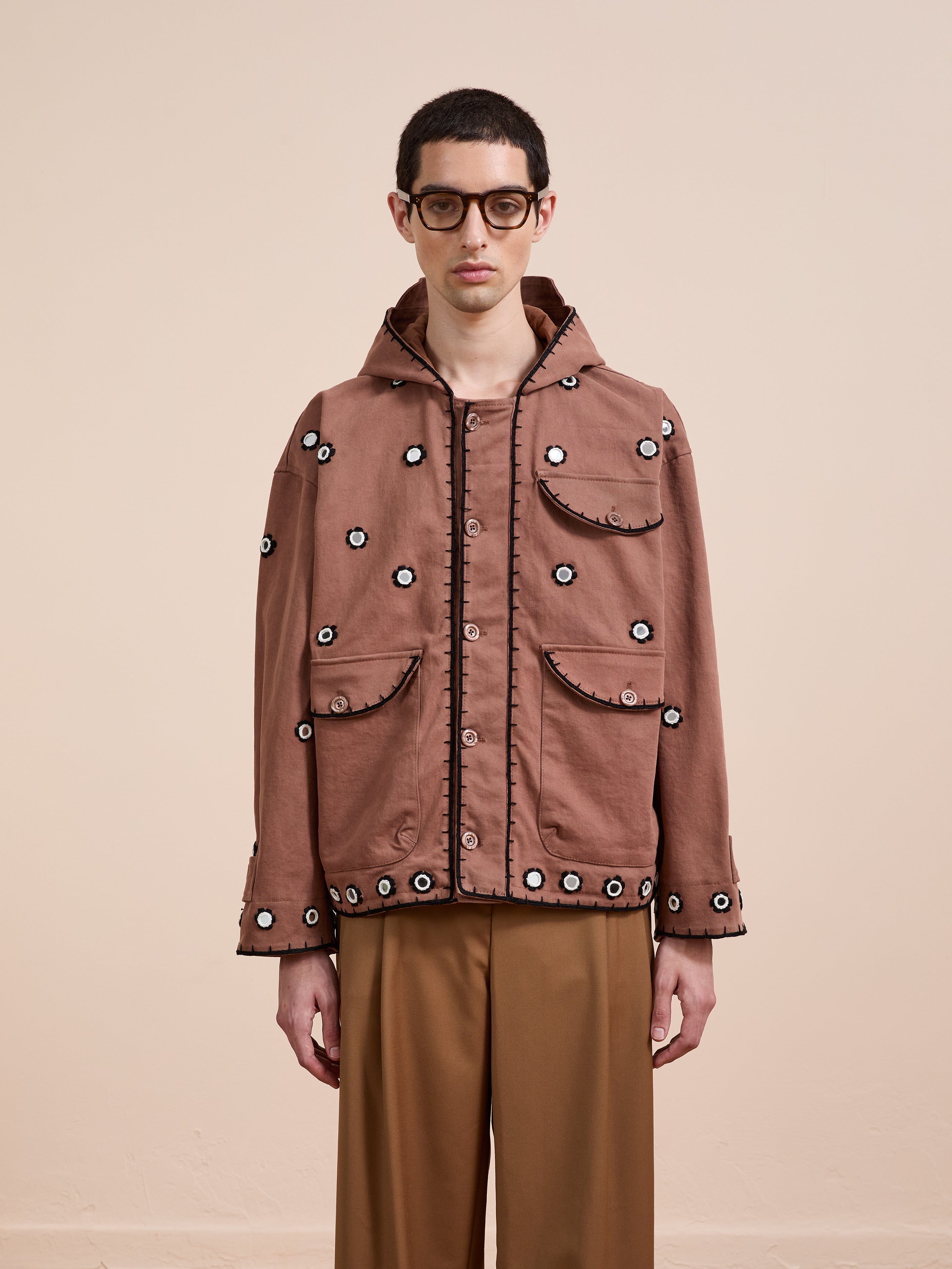 A person wearing glasses and the FOUND Hand Embroidered Hooded Mirror Anorak with sheesha-inspired patterns, paired with brown wide-leg trousers, stands against a plain beige background.