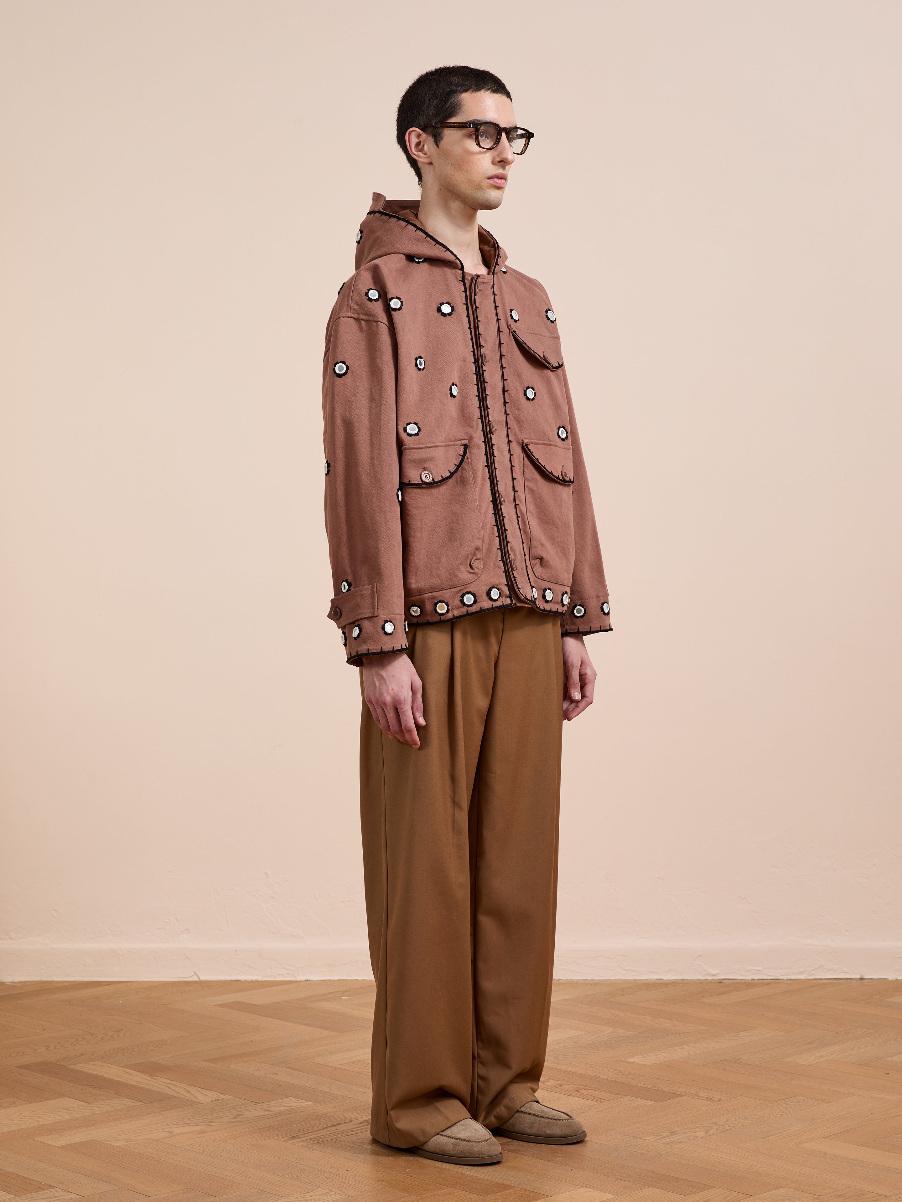 A person in profile wears glasses, FOUND's Hand Embroidered Hooded Mirror Anorak, wide brown trousers, and beige shoes on a wooden floor with a plain beige background.