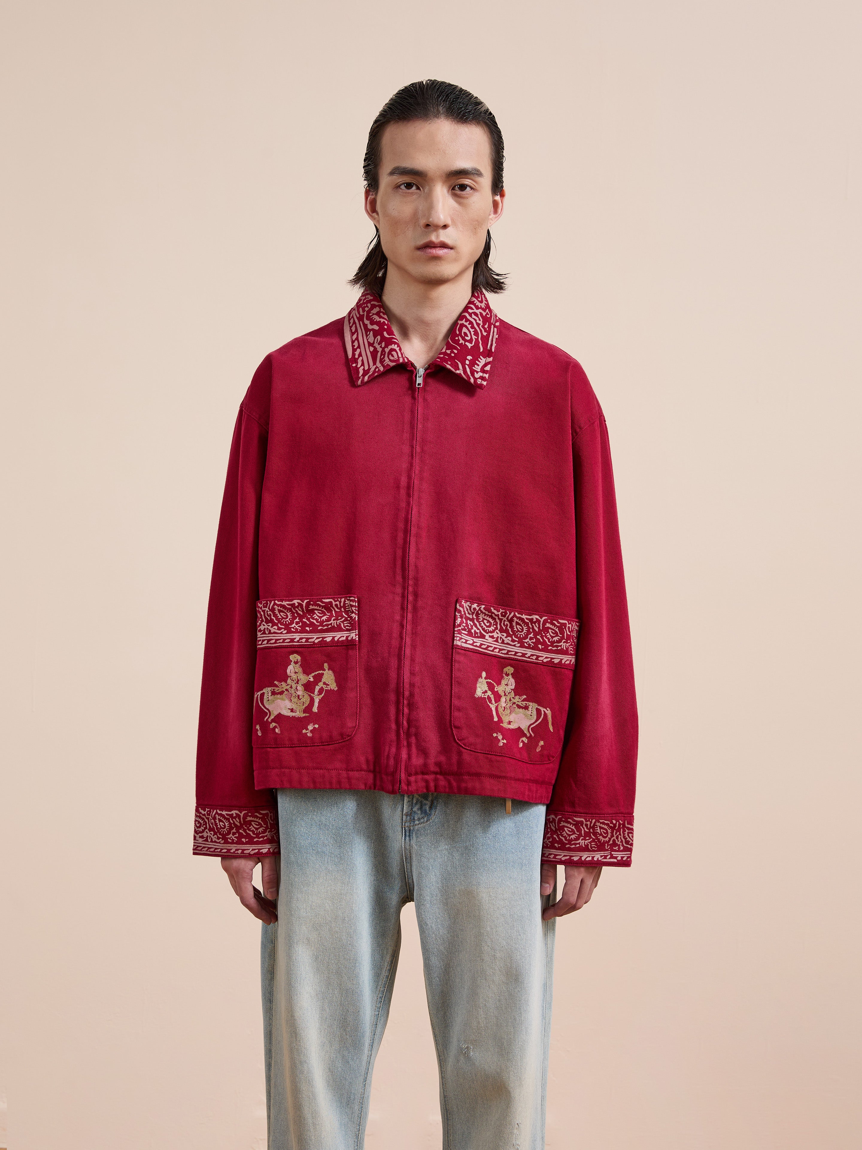 A person faces forward in the Found Abhala Printed Work Jacket, featuring equestrian motifs and horse images on the pockets, paired with light blue jeans against a plain beige background.