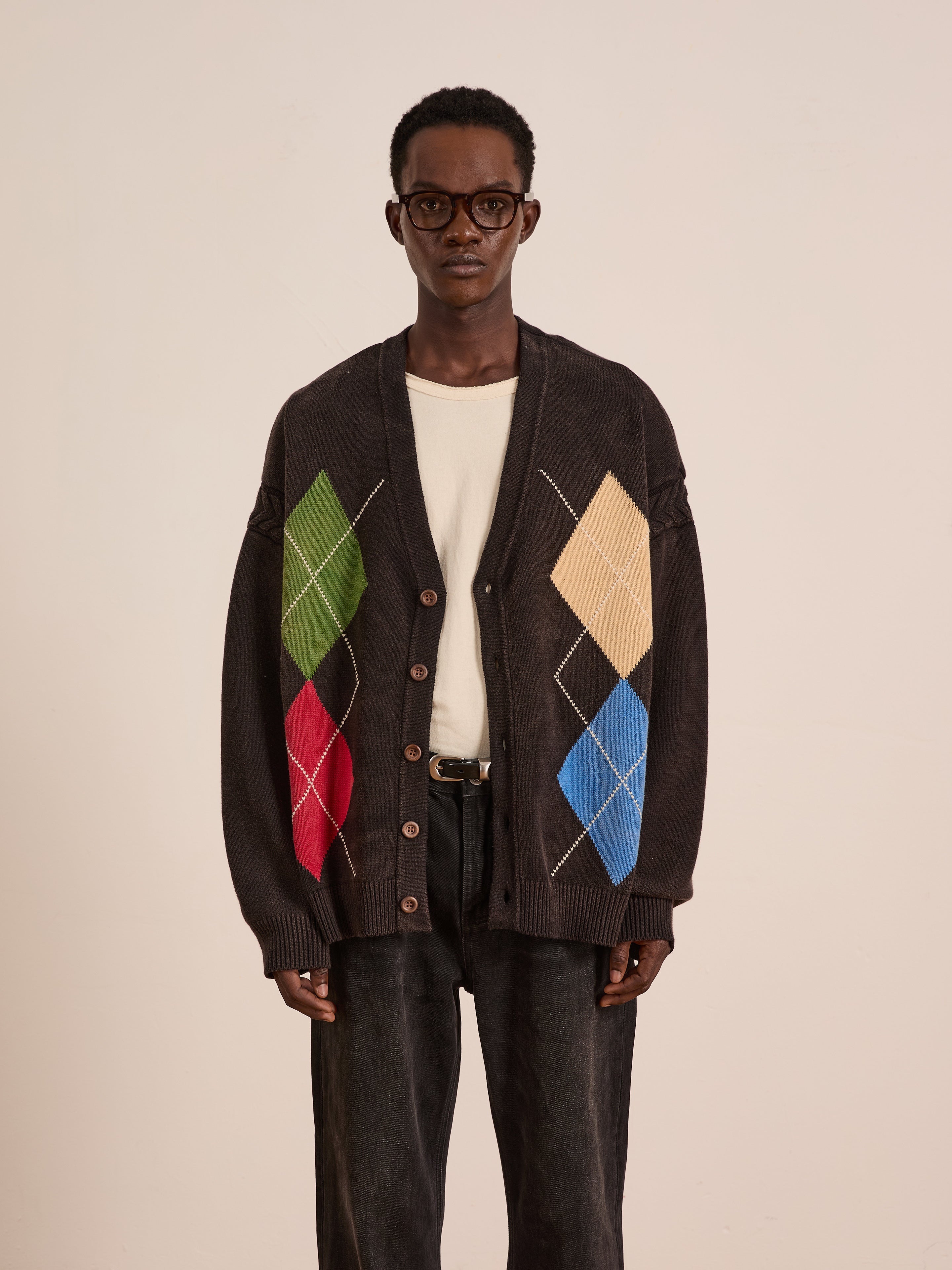 A person wearing glasses, a white t-shirt, dark pants, and a colorful FOUND Argyle Knit Cardigan stands against a plain light background.