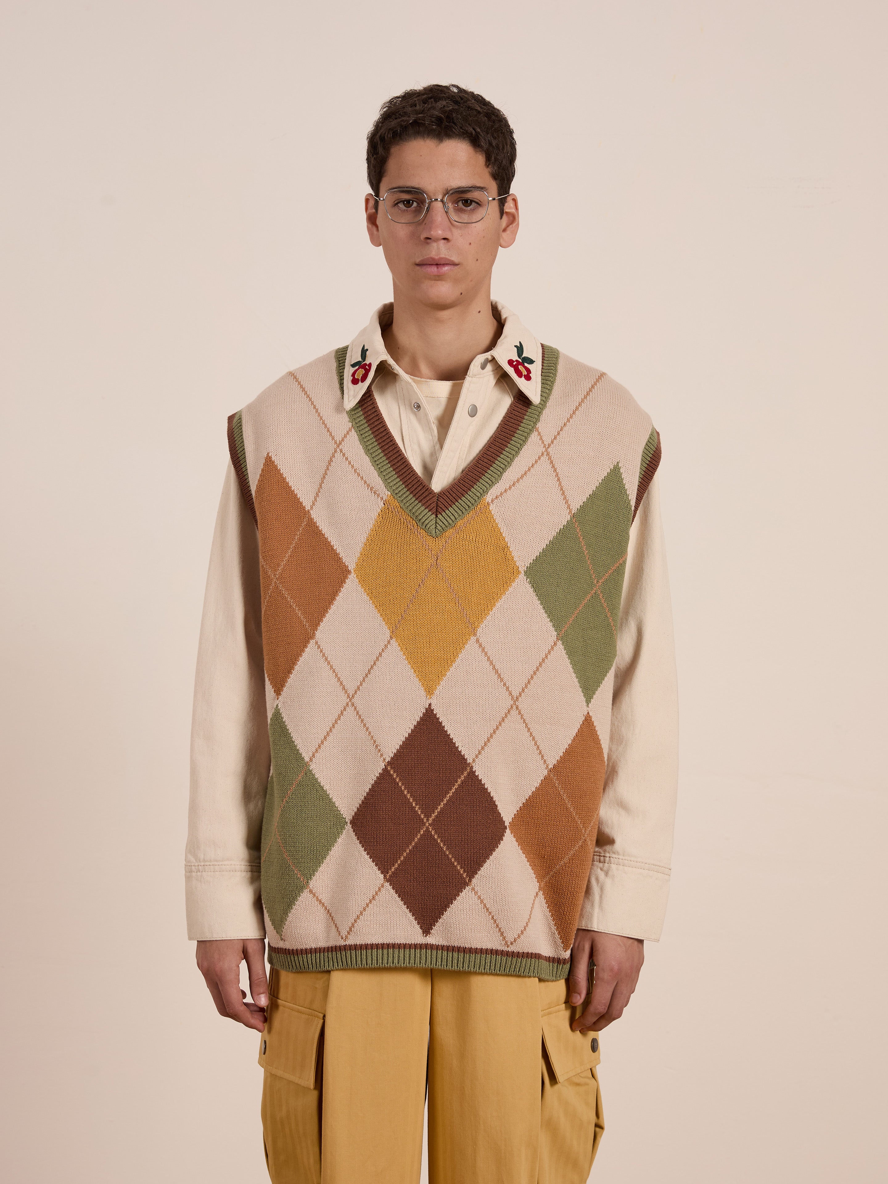 A person stands against a plain background, wearing glasses and showcasing the FOUND Argyle Knit Vest with a vintage-inspired diamond pattern over a beige floral embroidered shirt, paired with tan cargo pants—perfecting the unisex relaxed fit style.