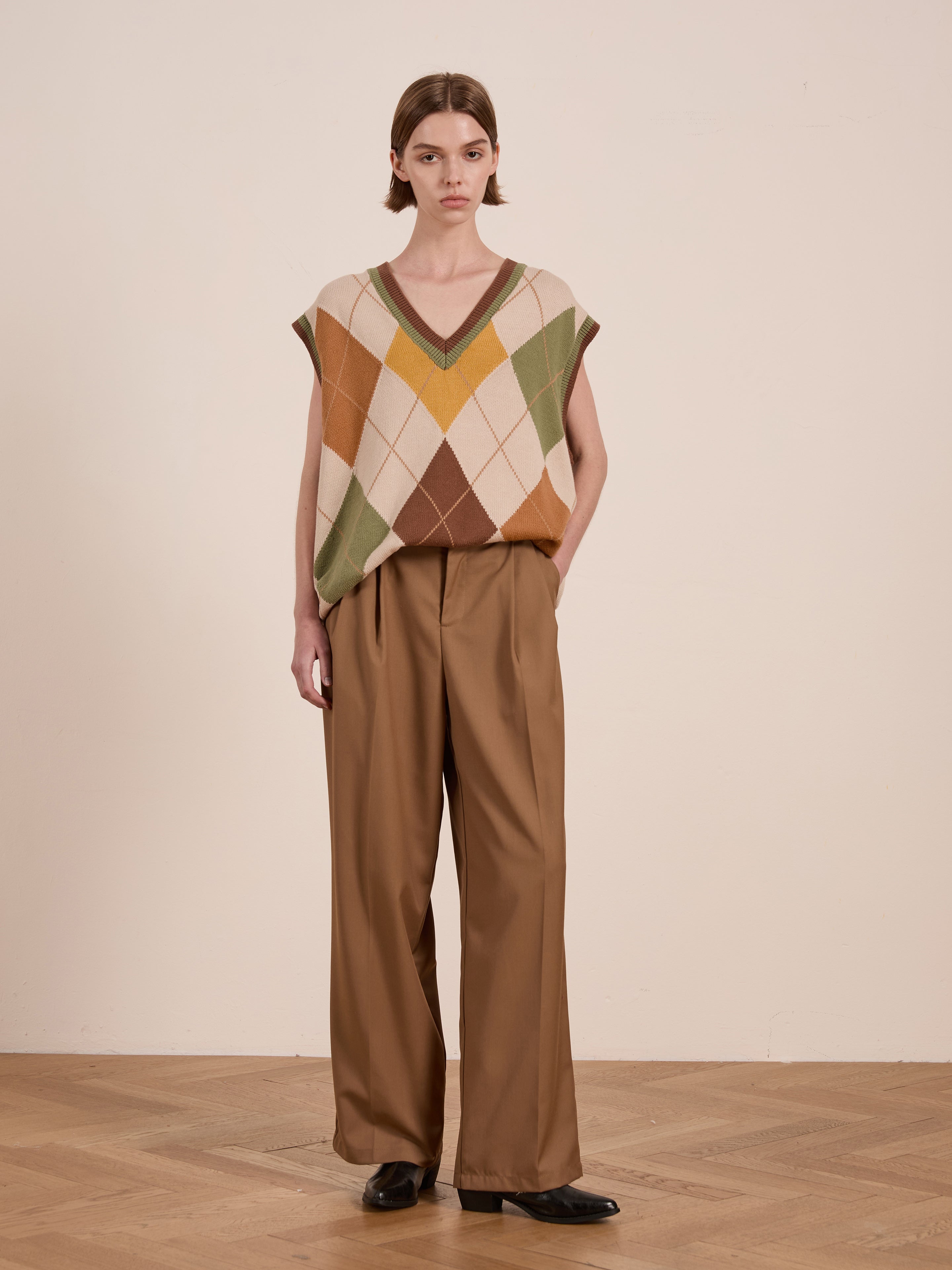 A person stands on a wooden floor wearing FOUND's vintage-inspired Argyle Knit Vest in a diamond pattern, paired with brown wide-leg trousers and black shoes. The unisex relaxed fit enhances the plain background beautifully.