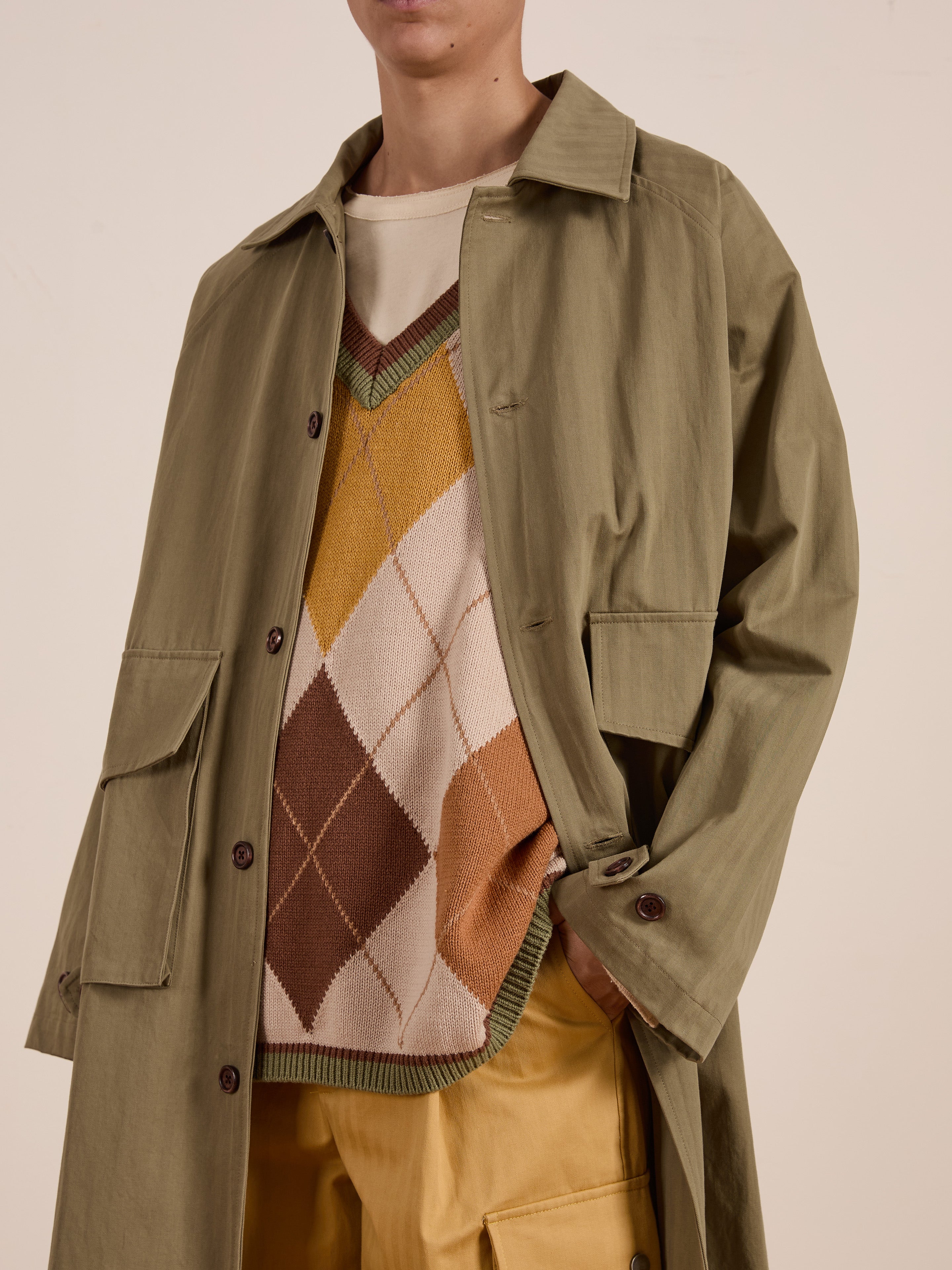 A person in a green trench coat pairs the FOUND Argyle Knit Vest, featuring a vintage-inspired diamond pattern in beige, orange, and brown tones, with unisex relaxed fit mustard yellow trousers.