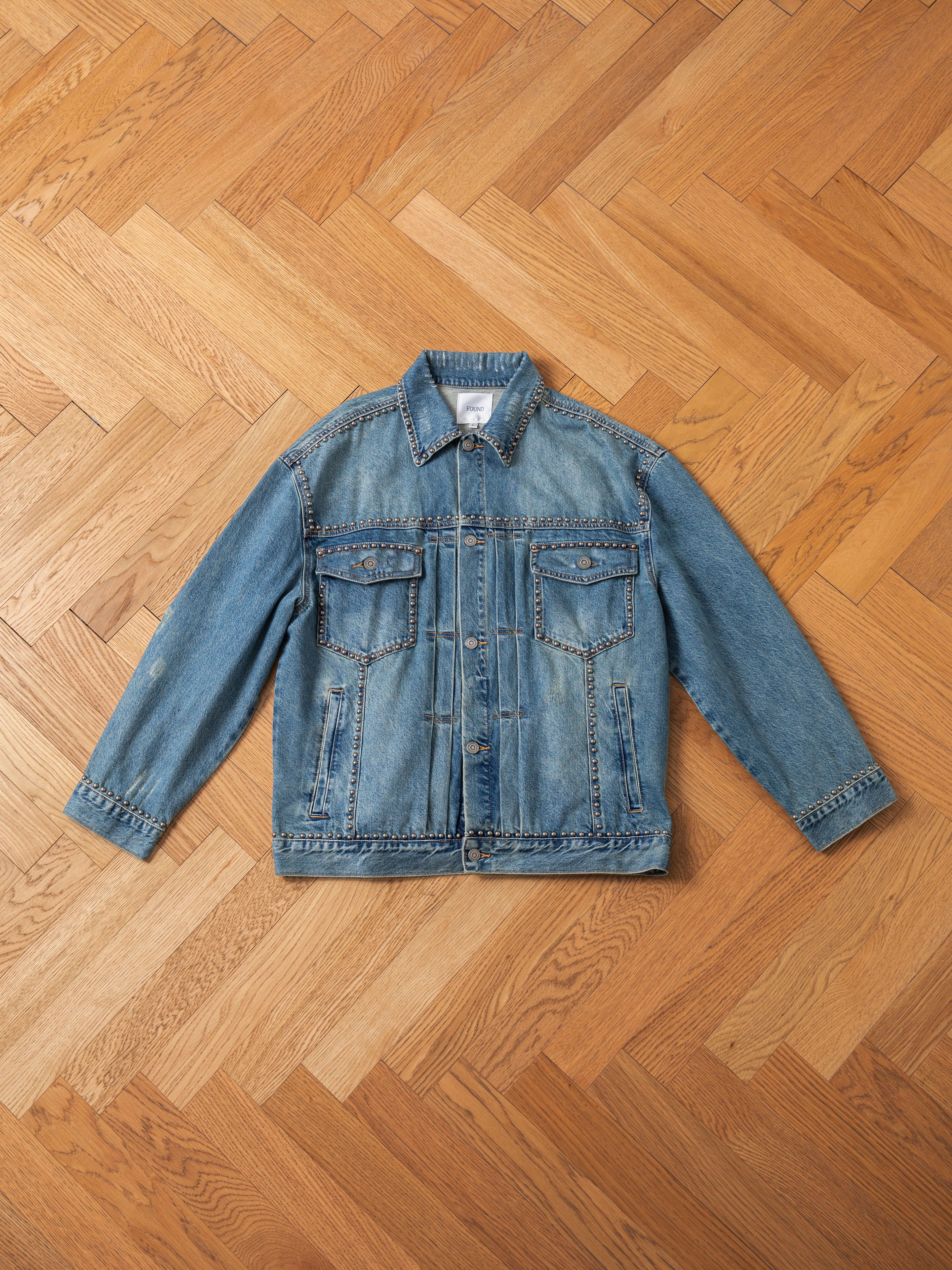 The FOUND Azar Studded Denim Trucker Jacket, featuring a button closure and two front chest pockets, showcases its vintage blue wash as it is laid flat on a wooden herringbone-pattern floor. Its oversized boxy fit adds an effortlessly cool vibe.