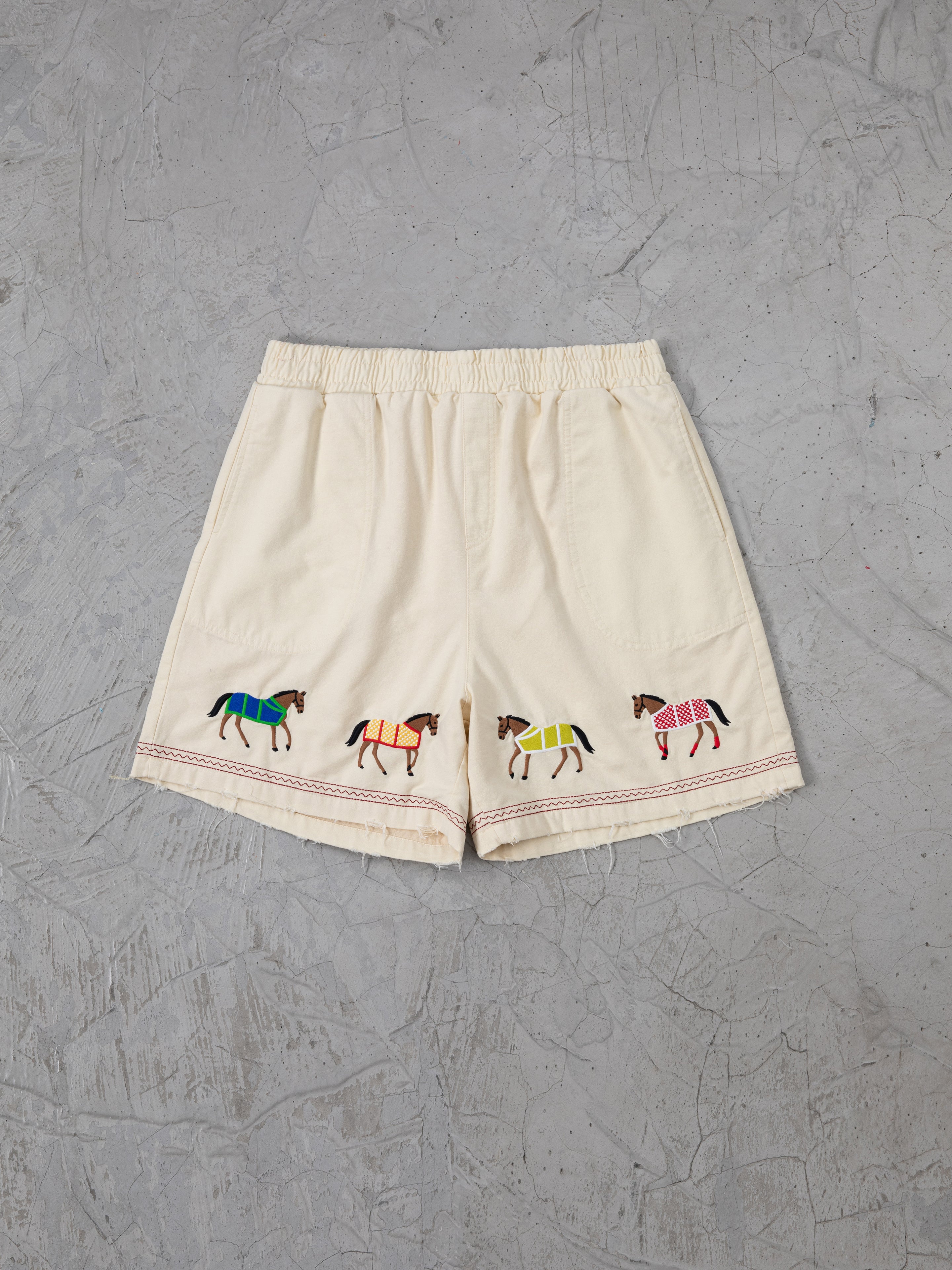 Profound's Embroidered Horse Shorts are designed in beige cotton twill with an elastic waistband, featuring vibrant embroidered horse motifs on a textured gray backdrop.