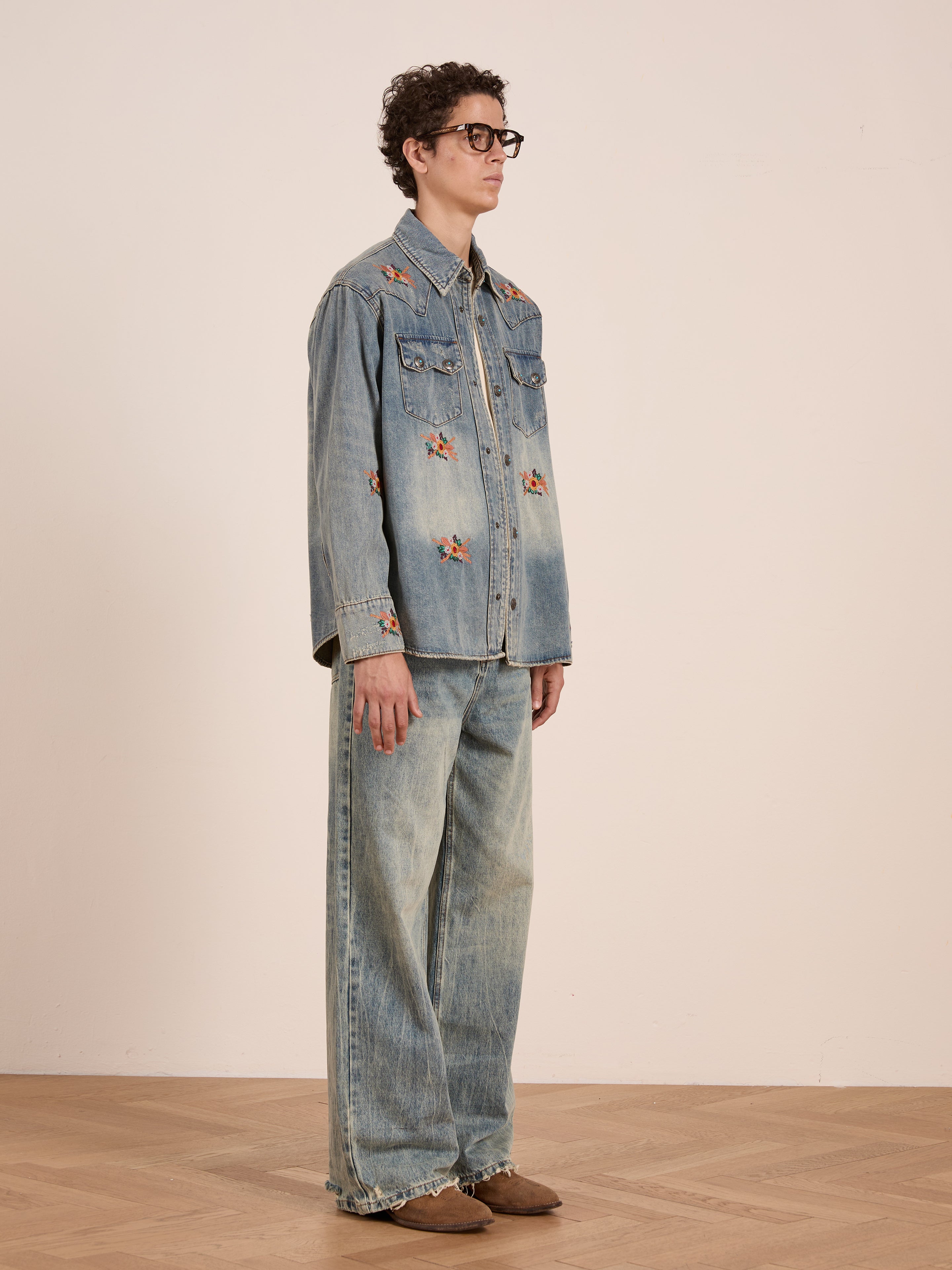 A person wearing the FOUND Bouquet Floral Embroidered Western Denim Shirt, matching relaxed wide-leg jeans, brown shoes, and glasses stands against a plain background.