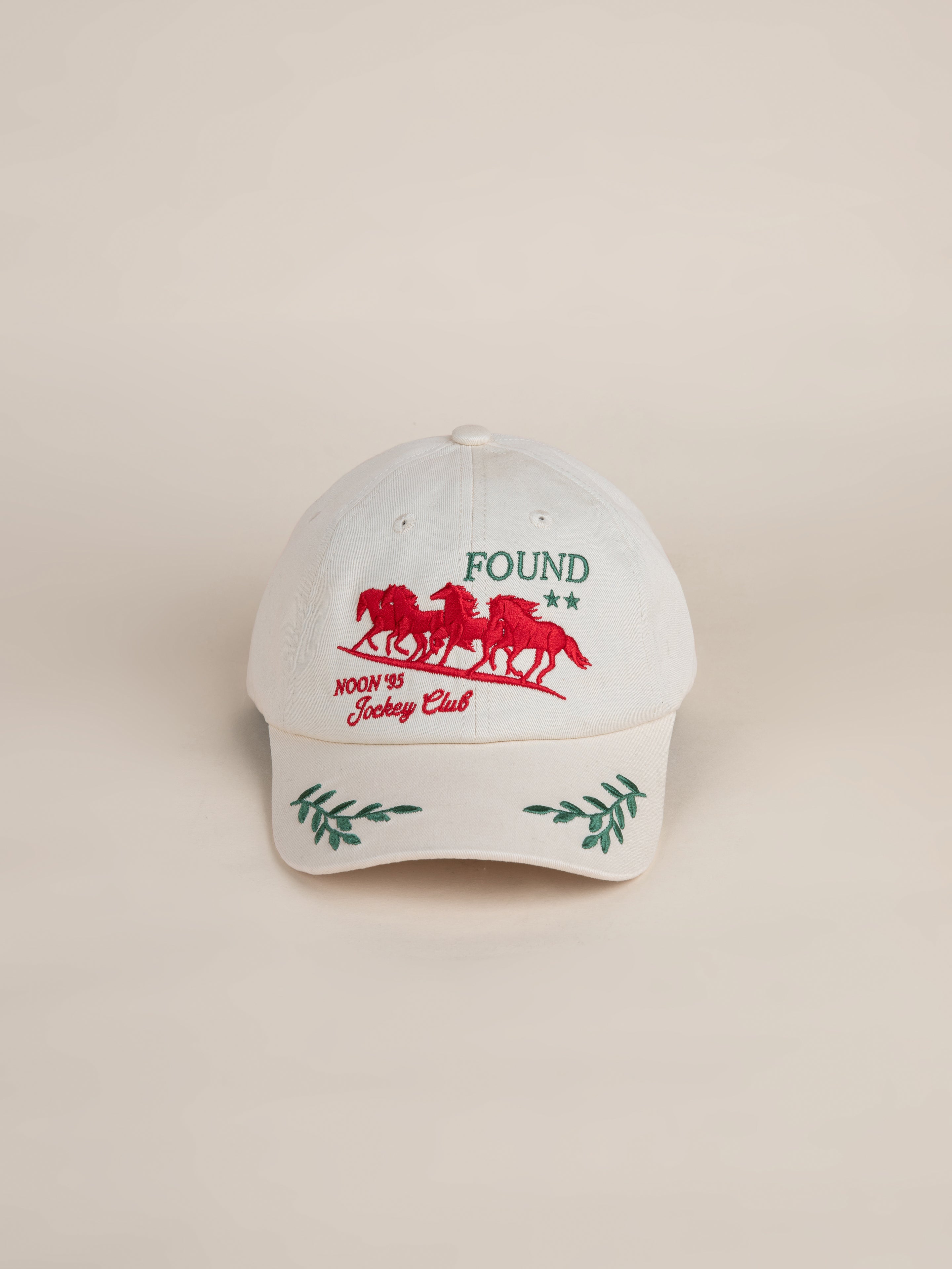 The Jockey Club Found Cap, by the brand FOUND, is a white baseball cap featuring red horse embroidery next to the logo and inscription "NOON '85 Jockey Club." The brim is adorned with green leaf designs, adding a natural flair to this stylish embroidered cap.