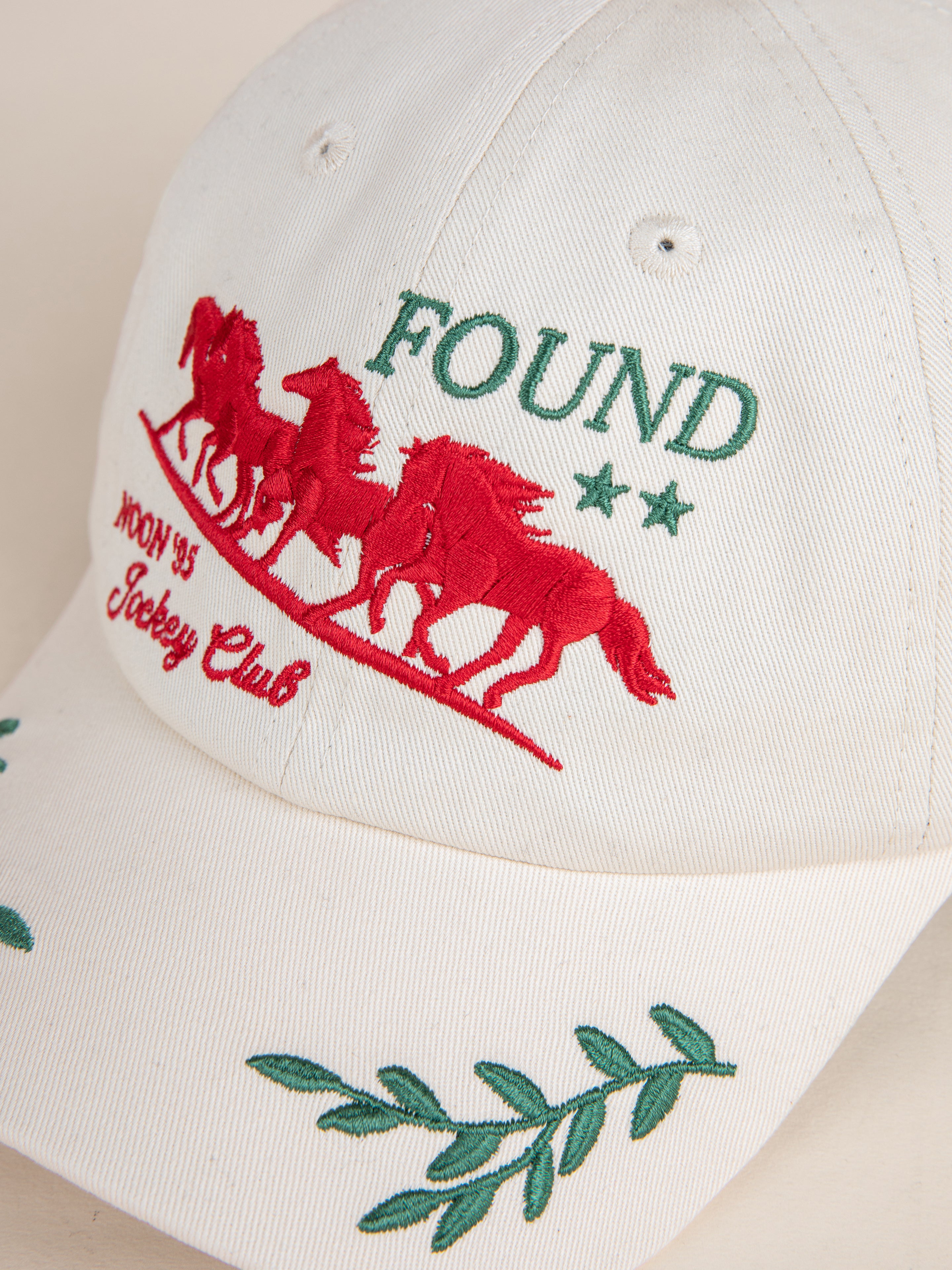 Jockey Club Found Cap by FOUND, showcasing a white embroidered design with the "FOUND" logo, accompanied by green "FOUND" text, two stars, red horse graphics, "MOON 93 Jockey Club" text, and lively green leaf decorations.