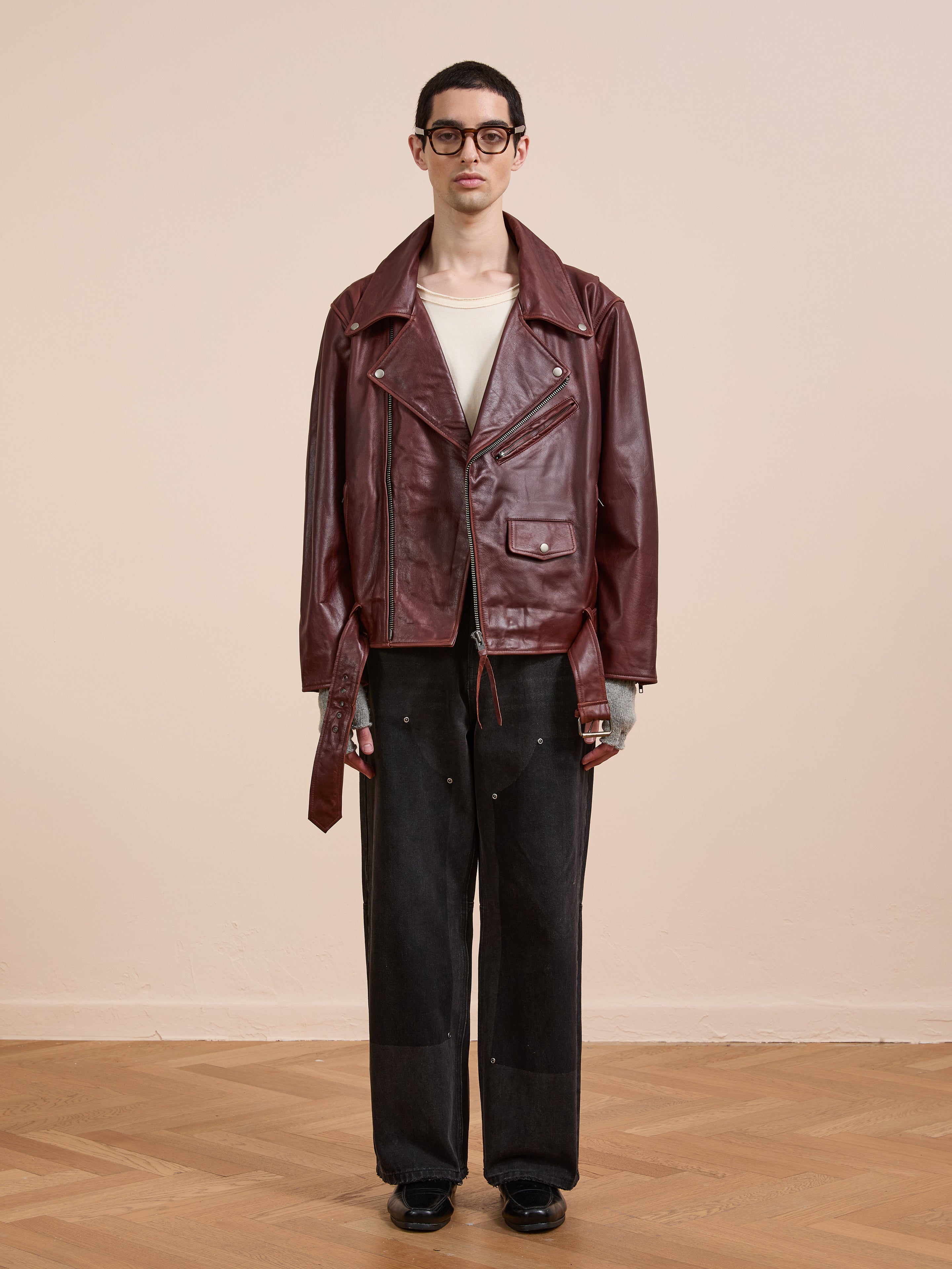 A person stands indoors against a plain beige wall, wearing black glasses, the FOUND Leather Biker Jacket in maroon leather, a white shirt, dark wide-leg pants, and black shoes.