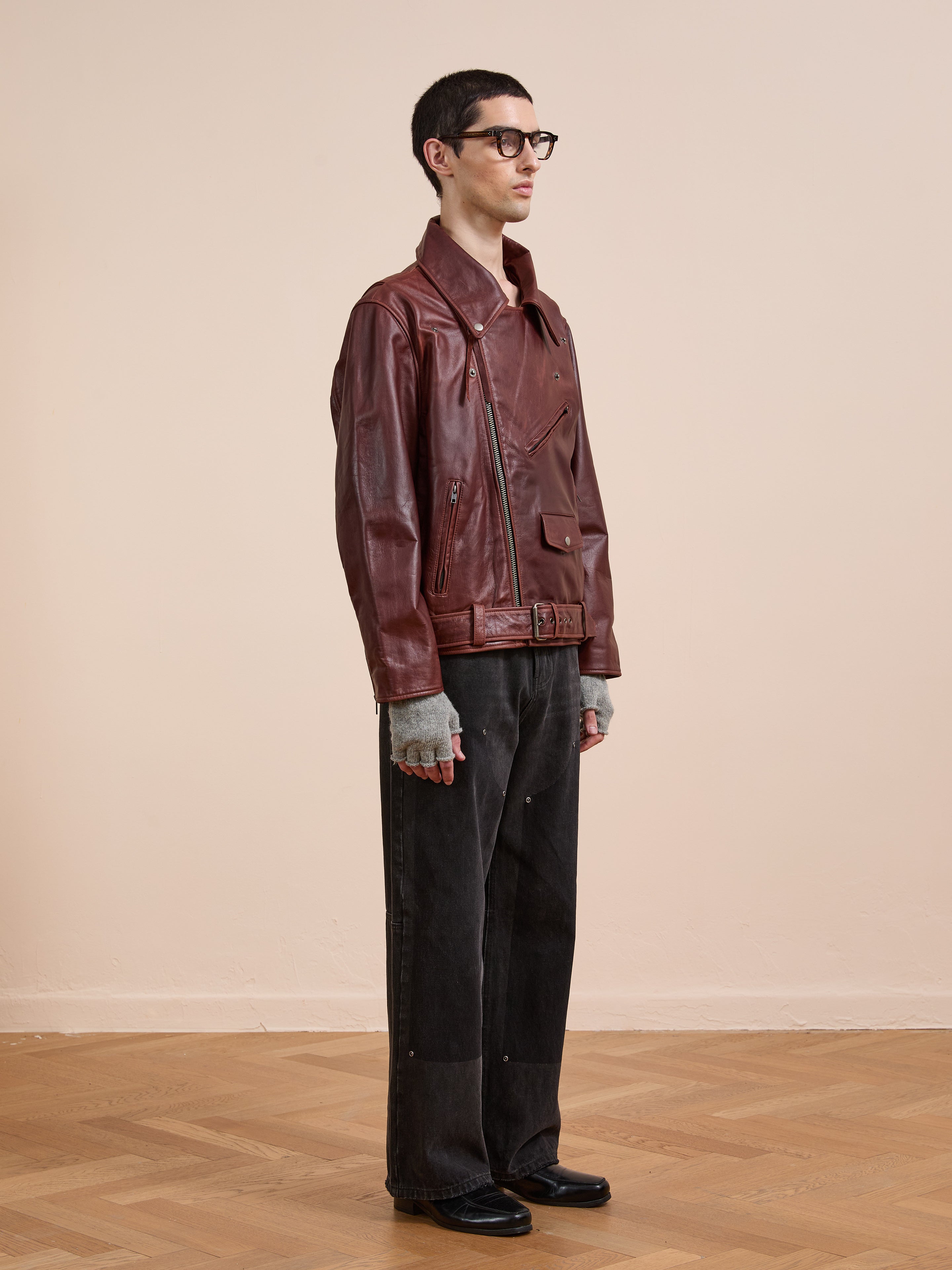 A person stands indoors on a wooden floor wearing FOUND’s Leather Biker Jacket, gray gloves, black wide-leg pants, black shoes, and glasses against a plain beige wall.