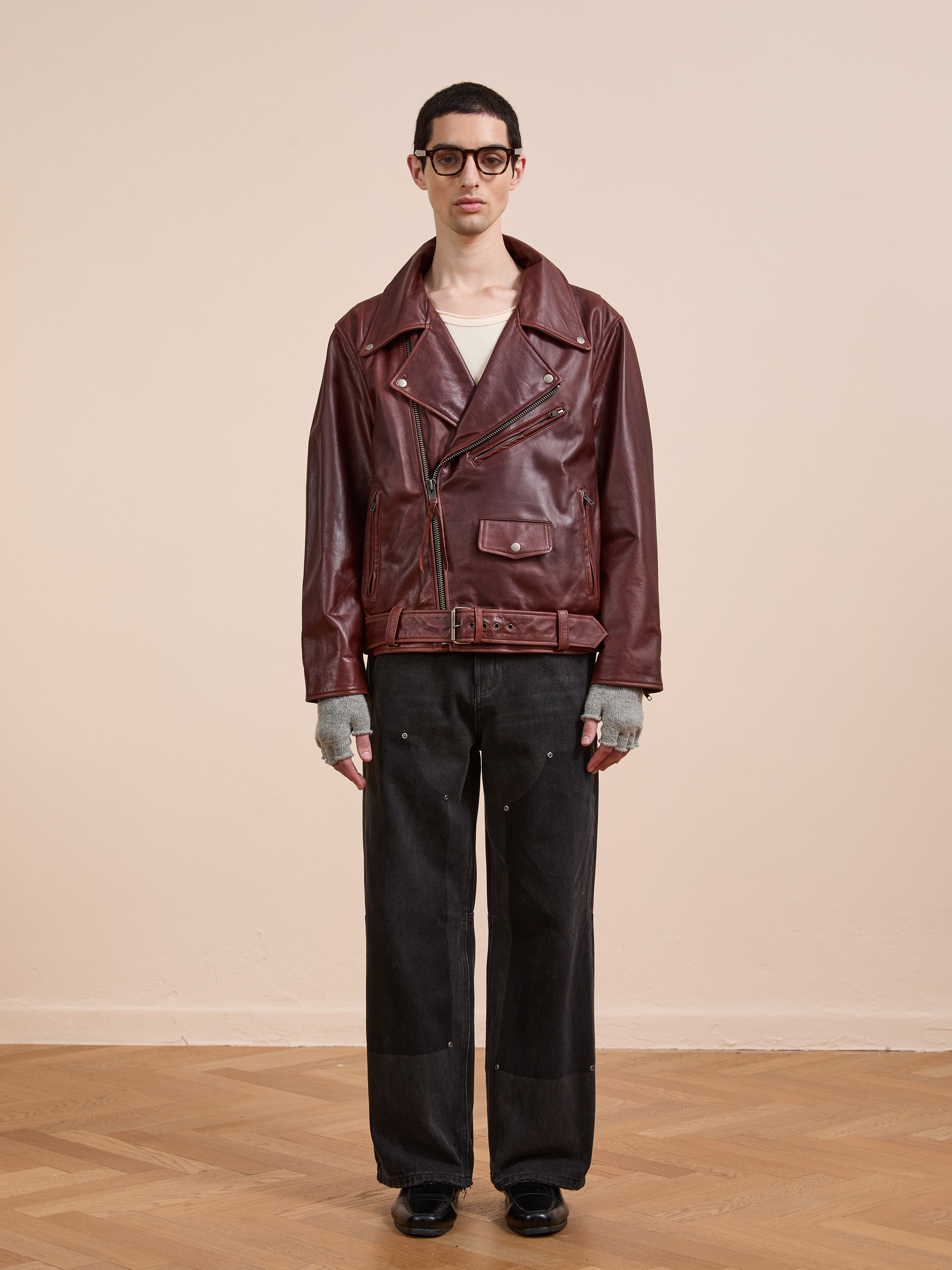 A person models the FOUND Leather Biker Jacket in maroon, paired with gray fingerless gloves, black wide-leg pants, and black shoes against a plain beige background.