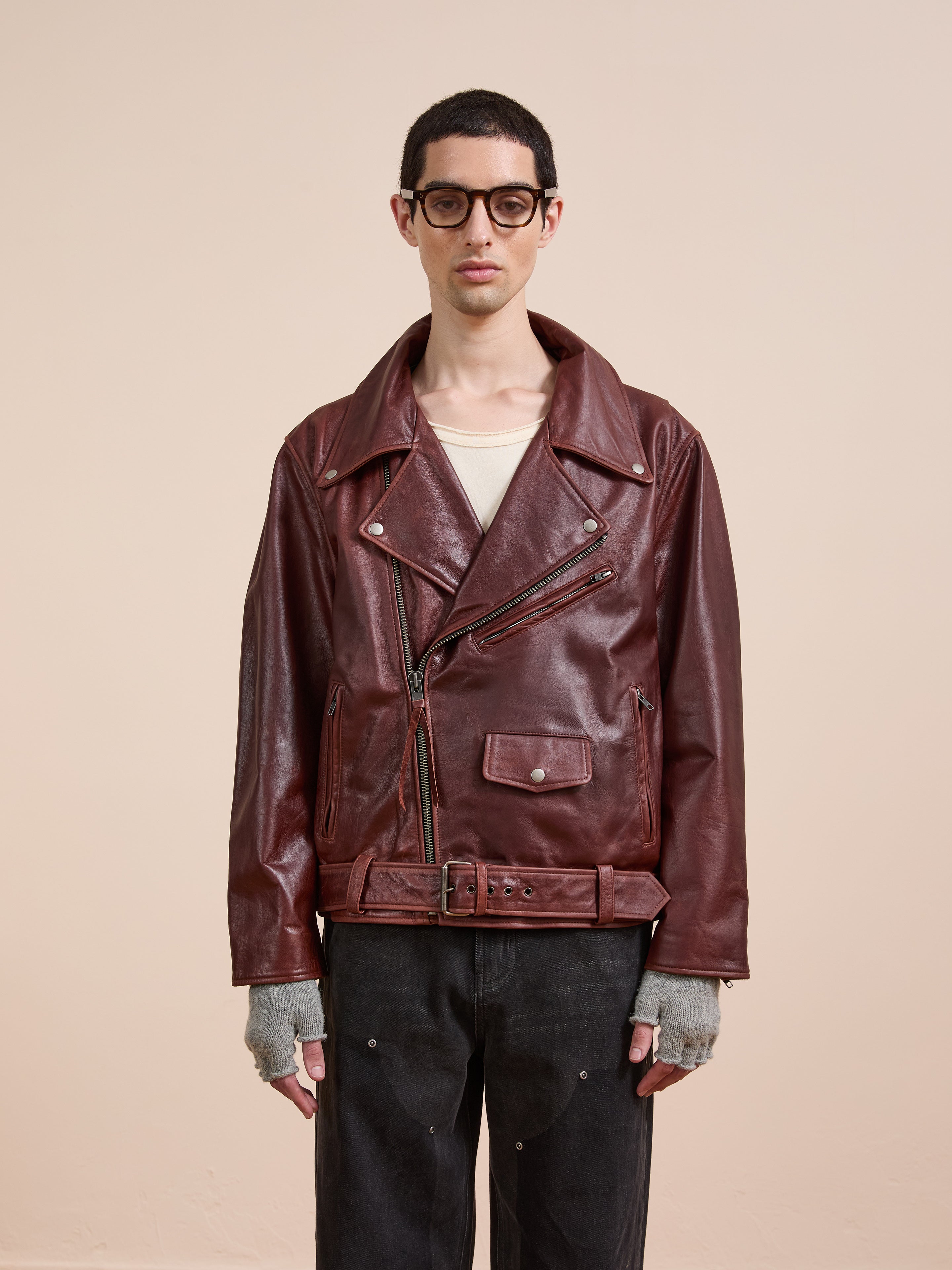 A person wearing FOUND’s burgundy Leather Biker Jacket, gray fingerless gloves, black jeans, and glasses stands against a plain beige background.