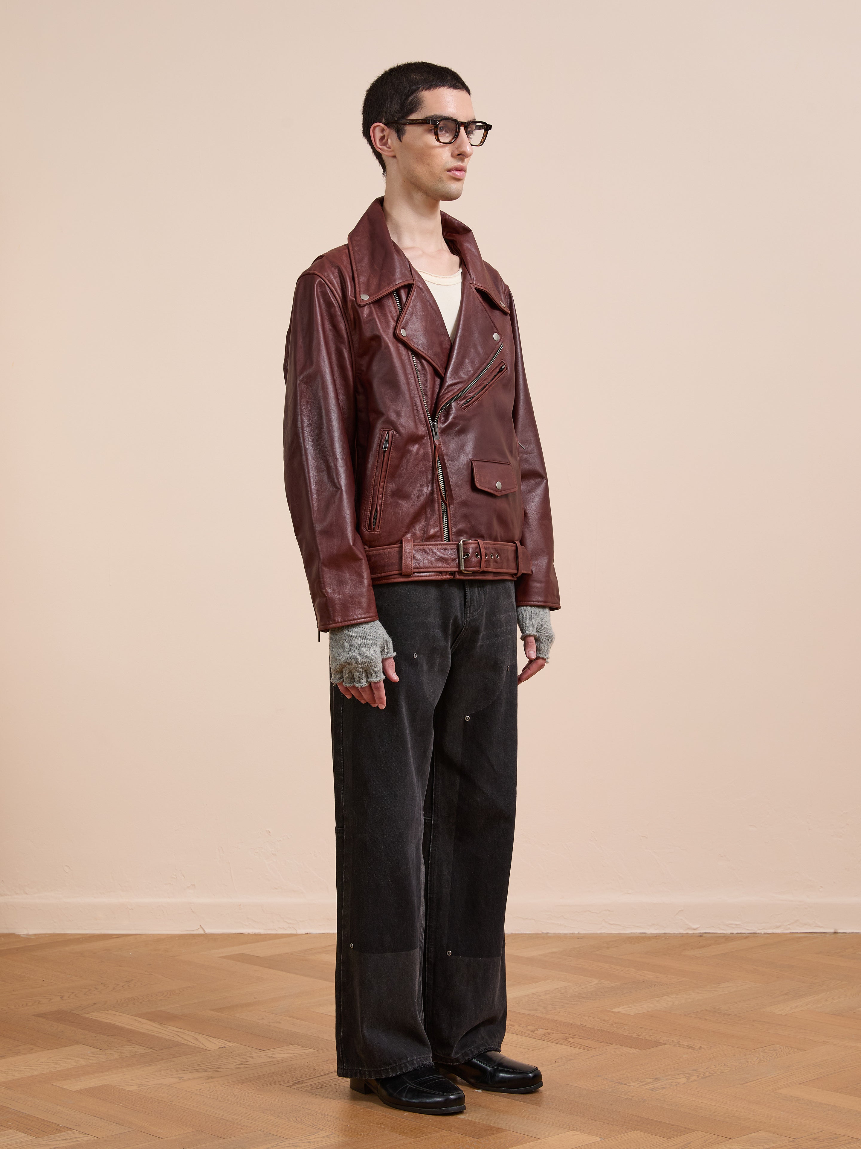 A person wearing the FOUND Leather Biker Jacket stands on a wooden floor against a plain beige wall, styled with gray fingerless gloves, black wide-leg pants, and black shoes.