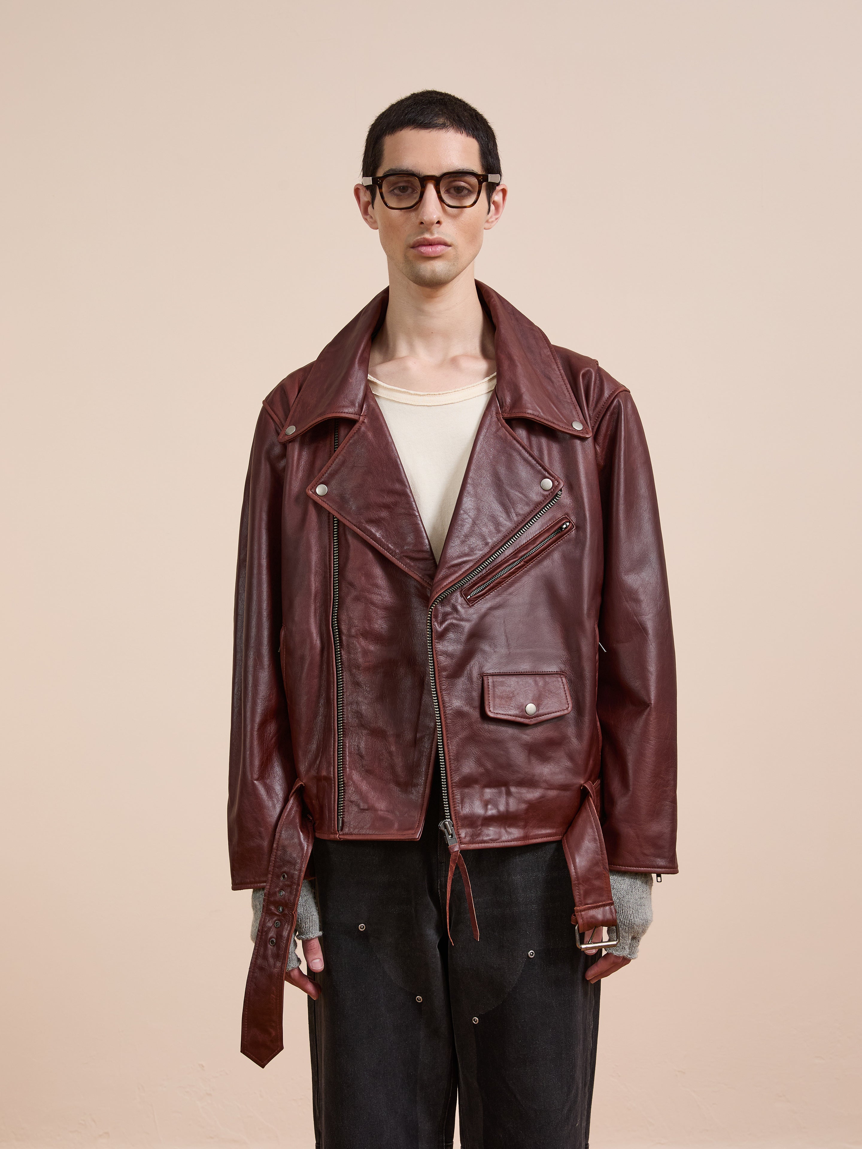 A person with short dark hair and glasses wears the FOUND Leather Biker Jacket over a beige shirt and dark pants, standing against a plain light-colored background.