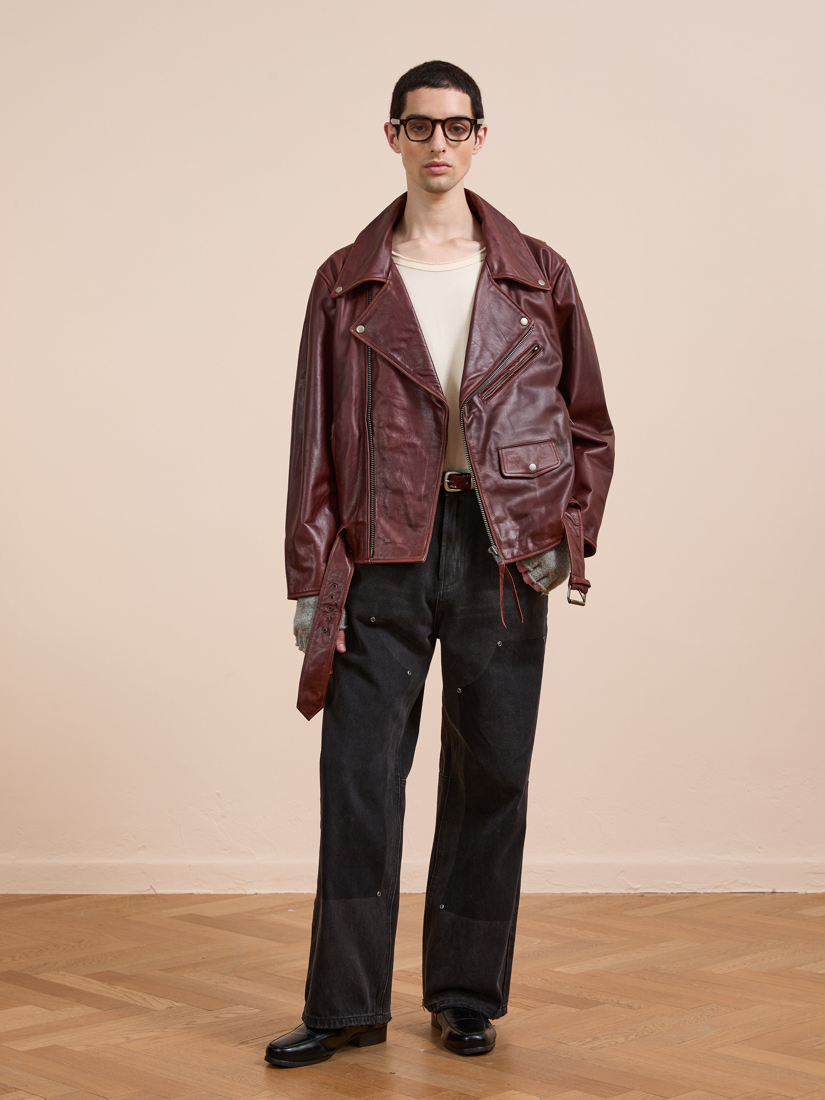 A person stands indoors against a beige wall and wooden floor, wearing the FOUND Leather Biker Jacket in maroon, paired with a white shirt, black wide-leg jeans, black shoes, and glasses.