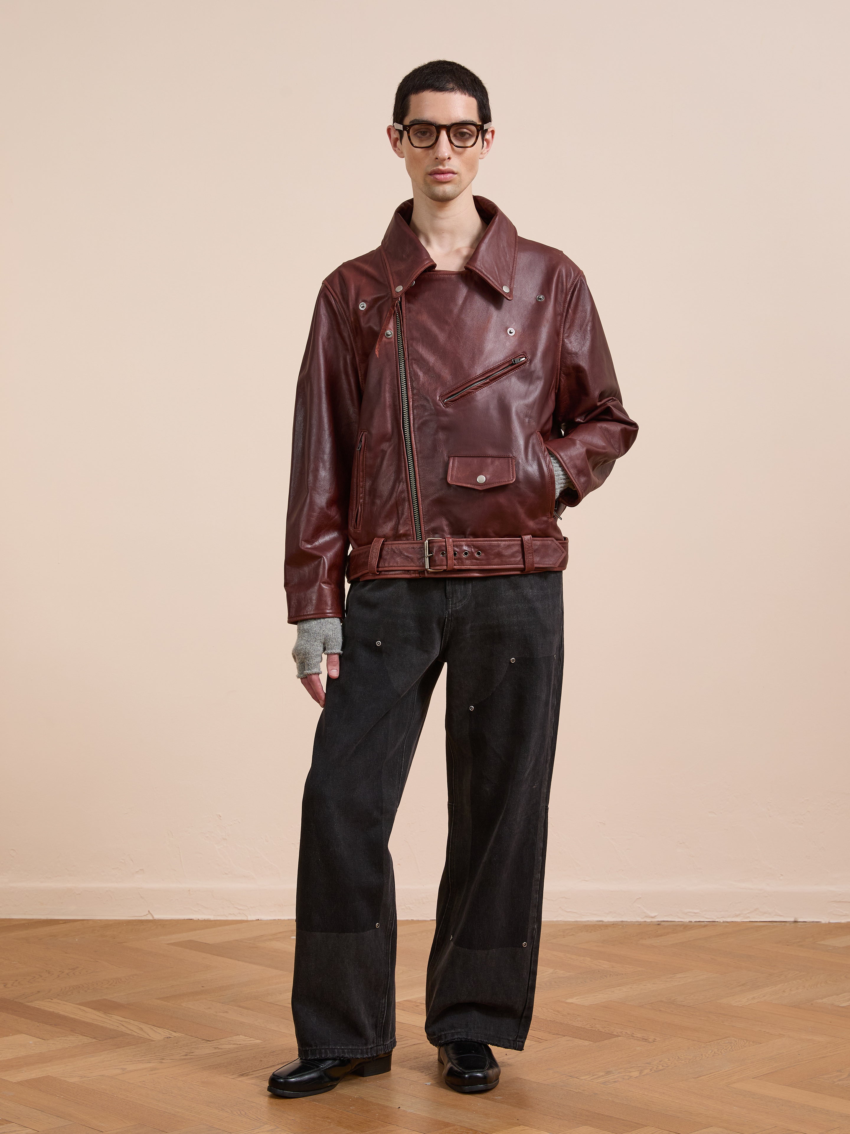Wearing a FOUND Leather Biker Jacket in burgundy, black wide-leg jeans, black shoes, gray fingerless gloves, and black glasses, a person stands on a hardwood floor against a plain beige wall.