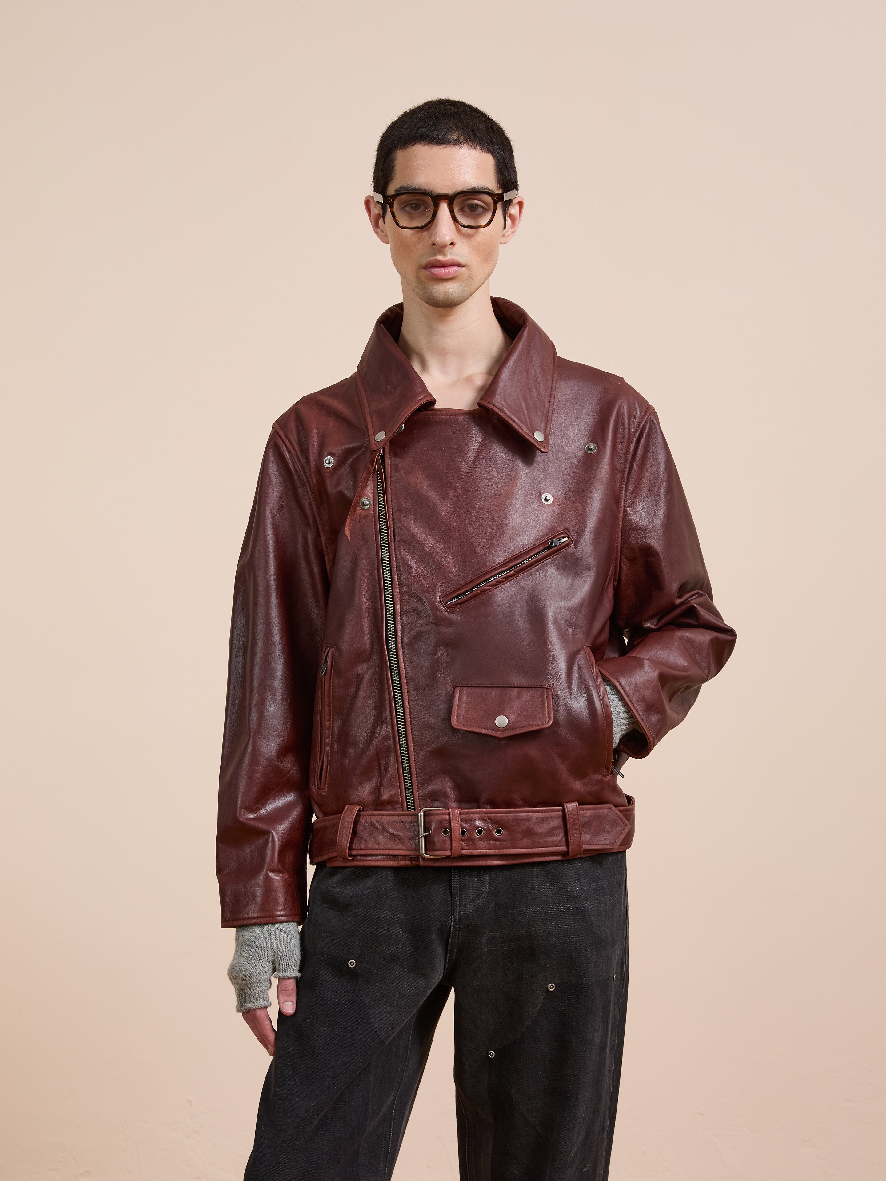 A person wearing glasses, the FOUND Leather Biker Jacket with an asymmetrical zipper in burgundy, and black pants stands against a plain beige background.