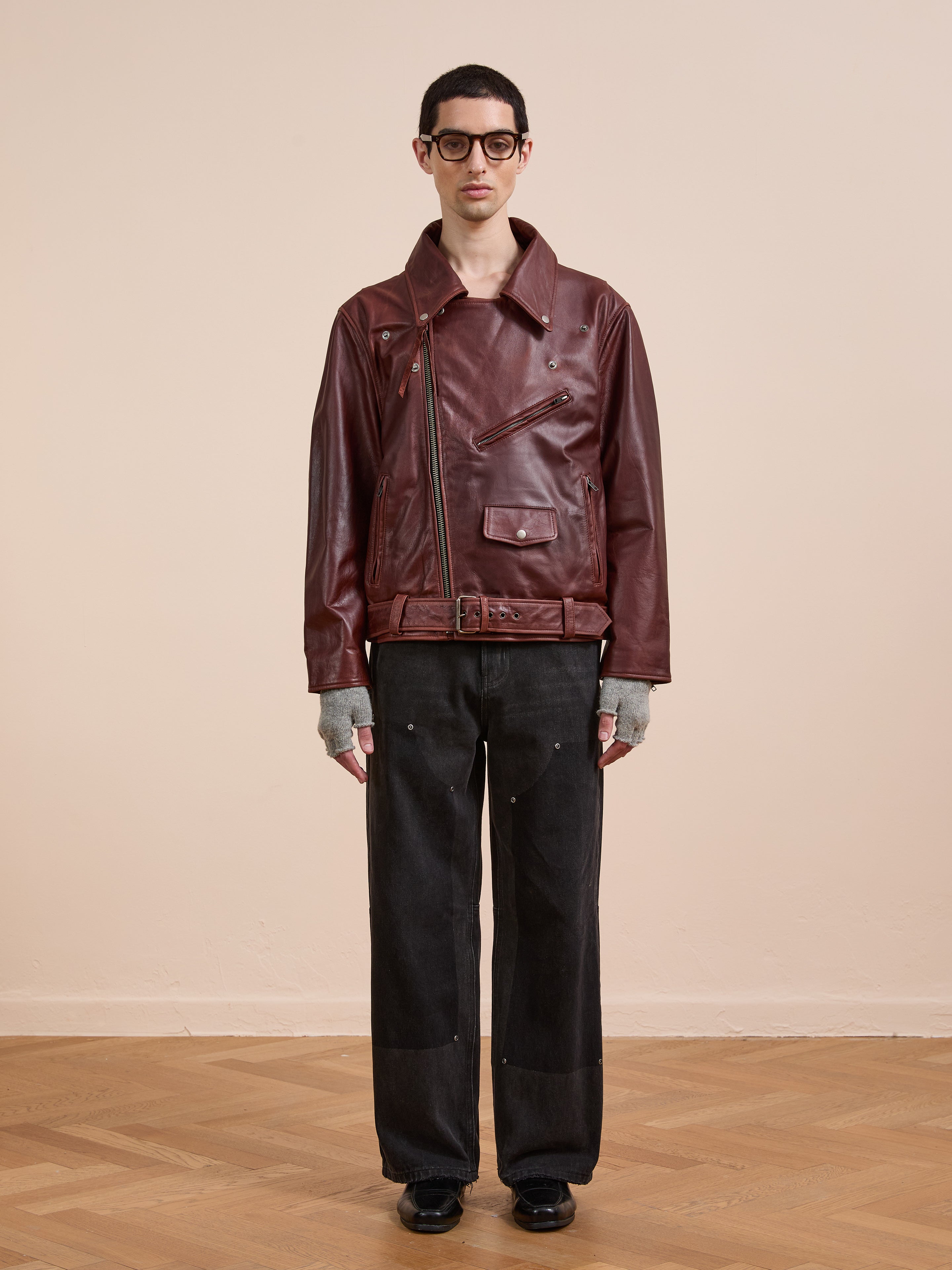 Someone wearing FOUND's maroon Leather Biker Jacket, black wide-leg pants, gray fingerless gloves, and black shoes stands against a plain beige wall on a wooden floor.