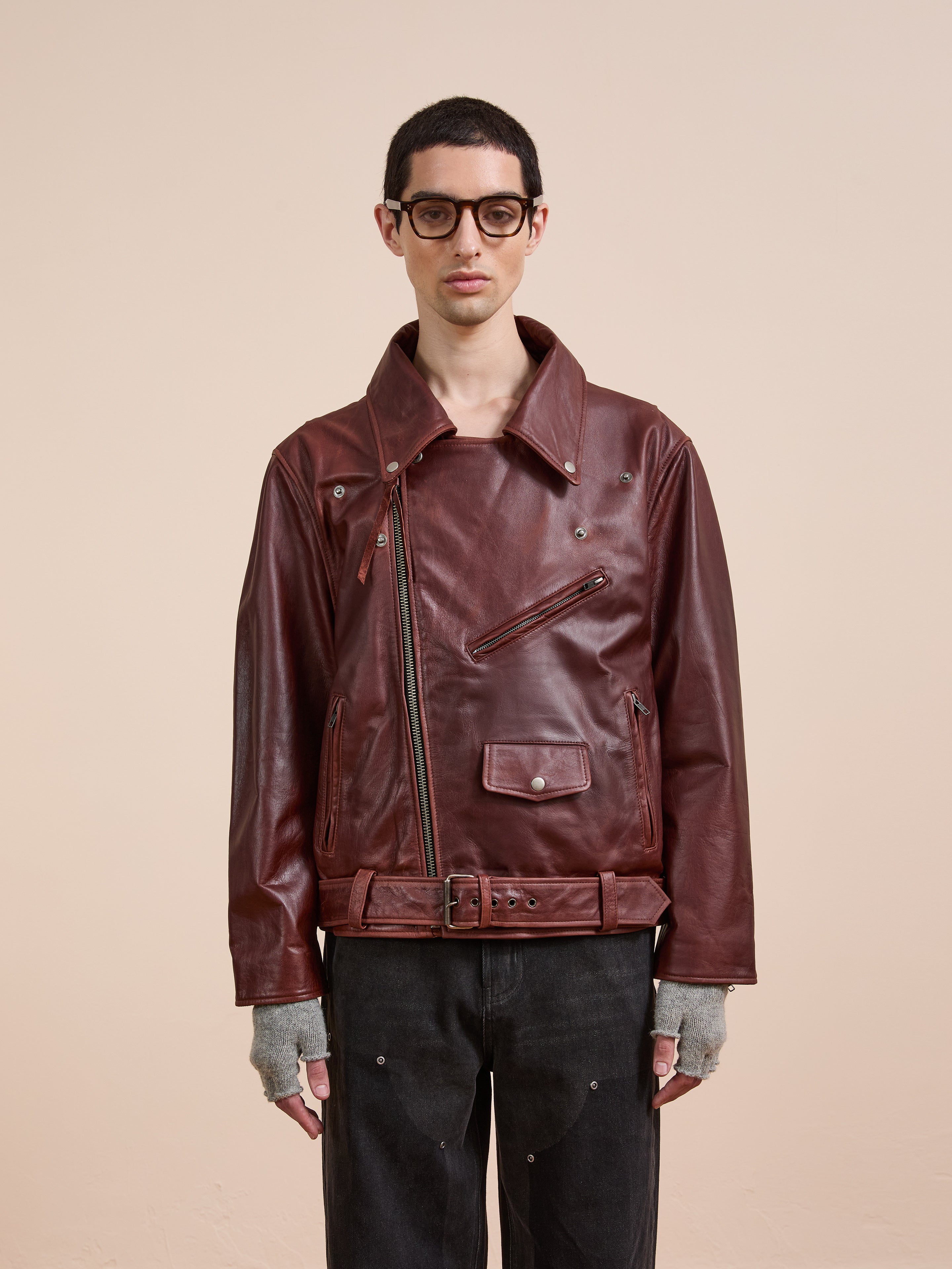 Someone stands against a plain beige background wearing FOUND’s Leather Biker Jacket in burgundy, paired with black pants, fingerless gray gloves, and large glasses.