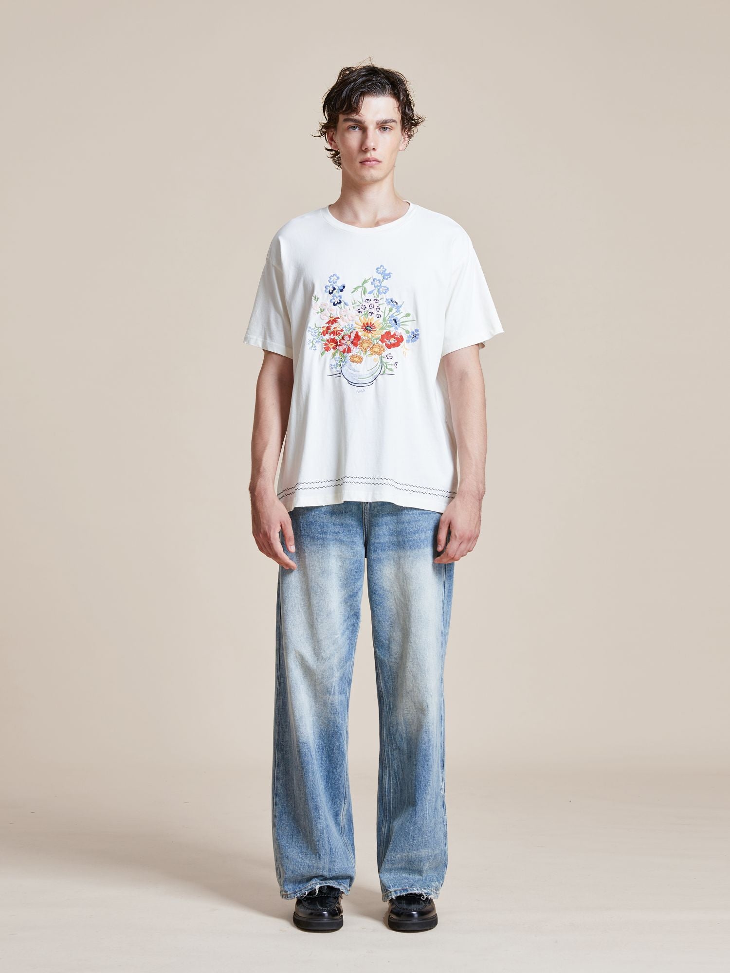 A person stands facing forward, wearing a Bouquet Flowers Tee from Found with traditional Punjab Phulkari embroidery and wide-leg blue jeans, against a plain beige background.