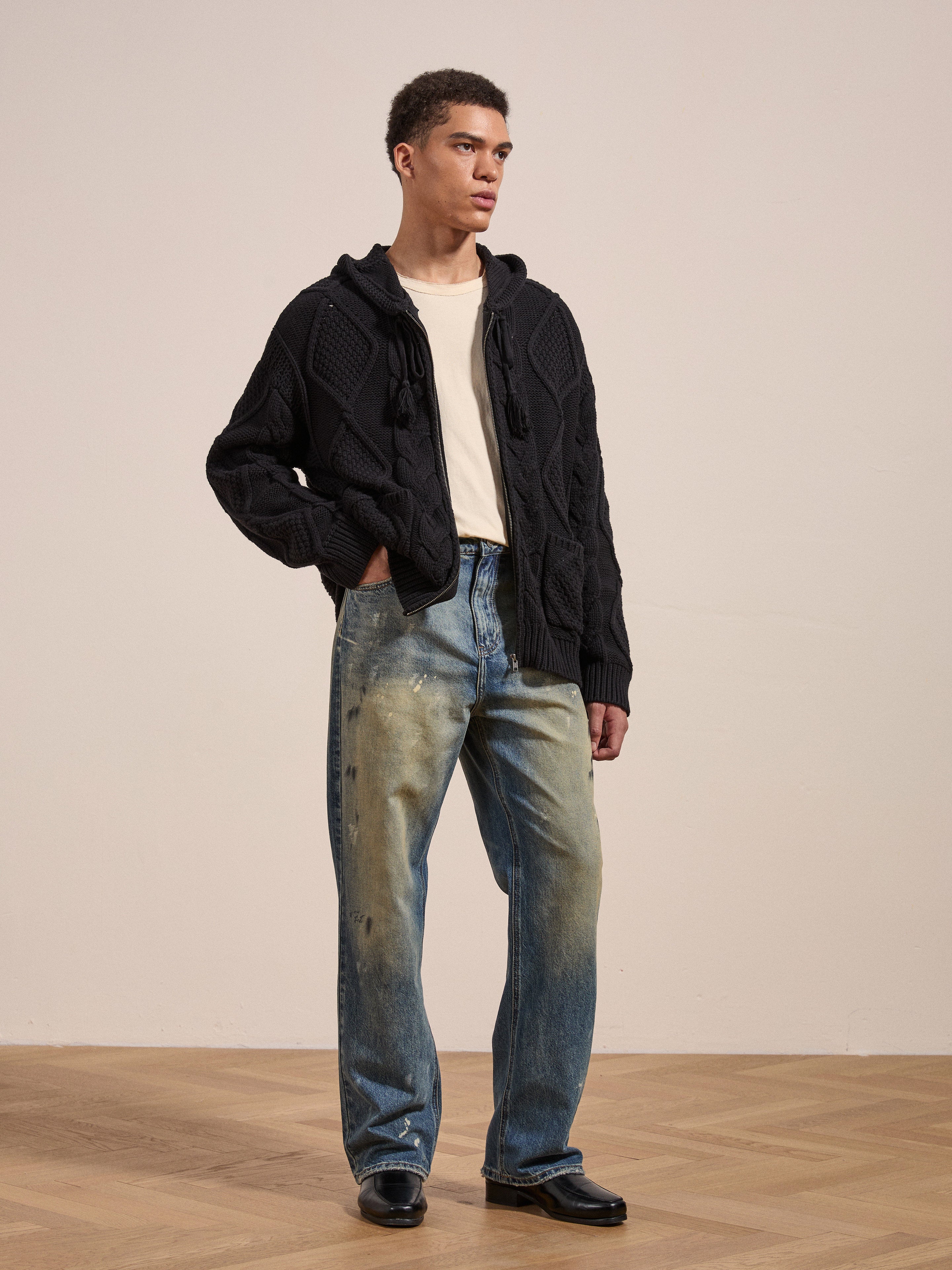 A person stands indoors on a wooden floor, dressed in Found's black Zip Up Cable Knit Hoodie, a white t-shirt, and worn blue jeans with black shoes. The heavyweight cotton blend yarn of the hoodie from Found provides a cozy look and feel.