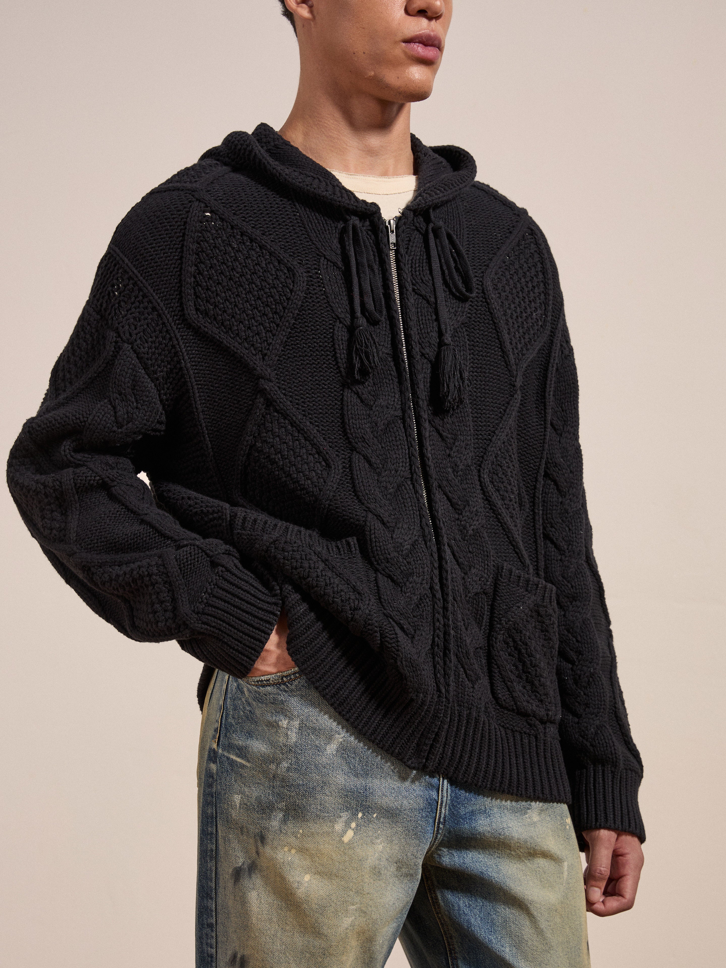 A person is wearing a black Found Zip Up Cable Knit Hoodie made of heavyweight cotton blend yarn, with front pockets and distressed blue jeans, standing against a plain background. The cozy look and feel of the outfit exudes comfort.