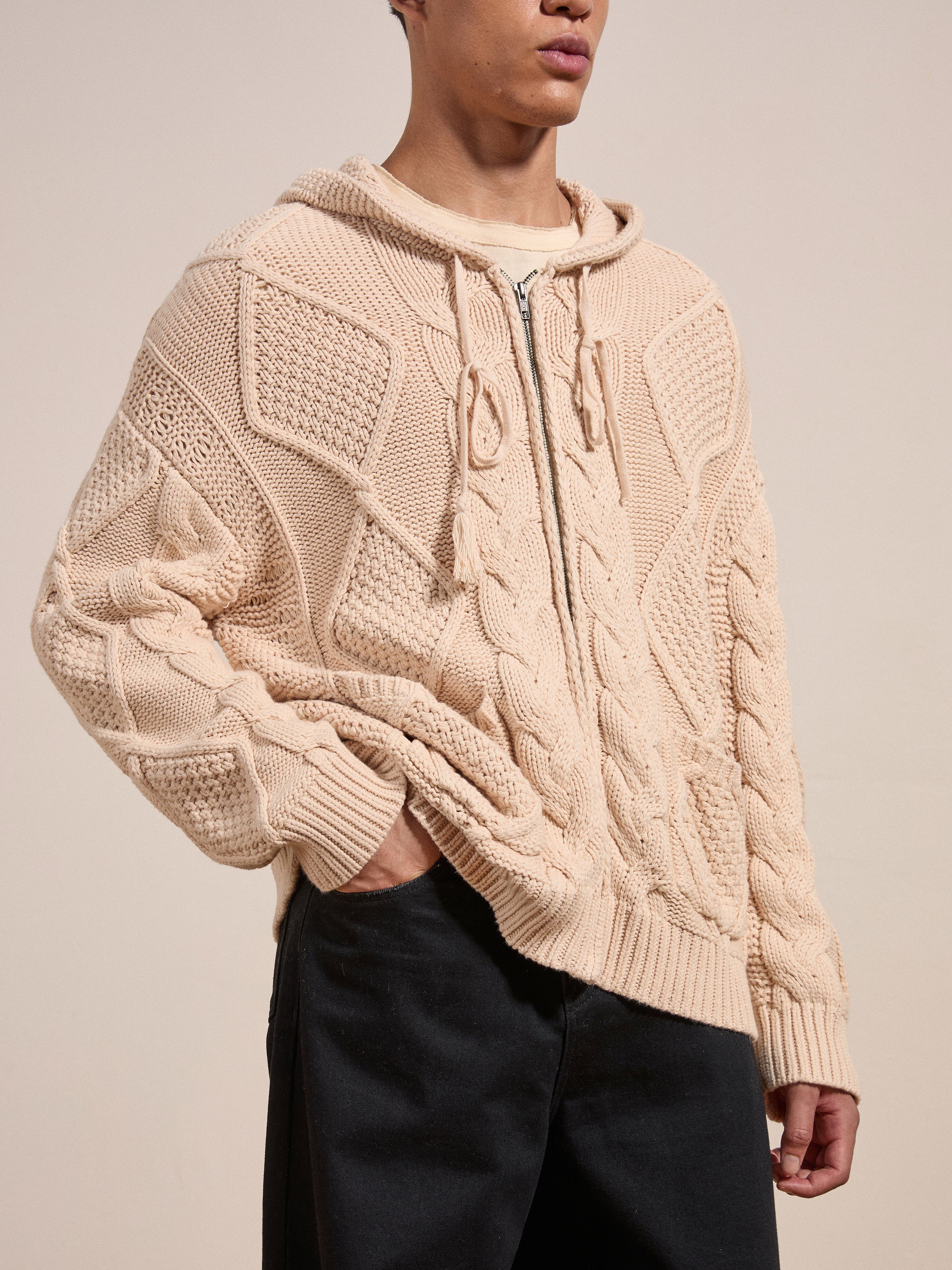 A person wearing a cozy beige Zip Up Cable Knit Hoodie by Found, crafted from a heavyweight cotton blend, stands with one hand in their pocket.