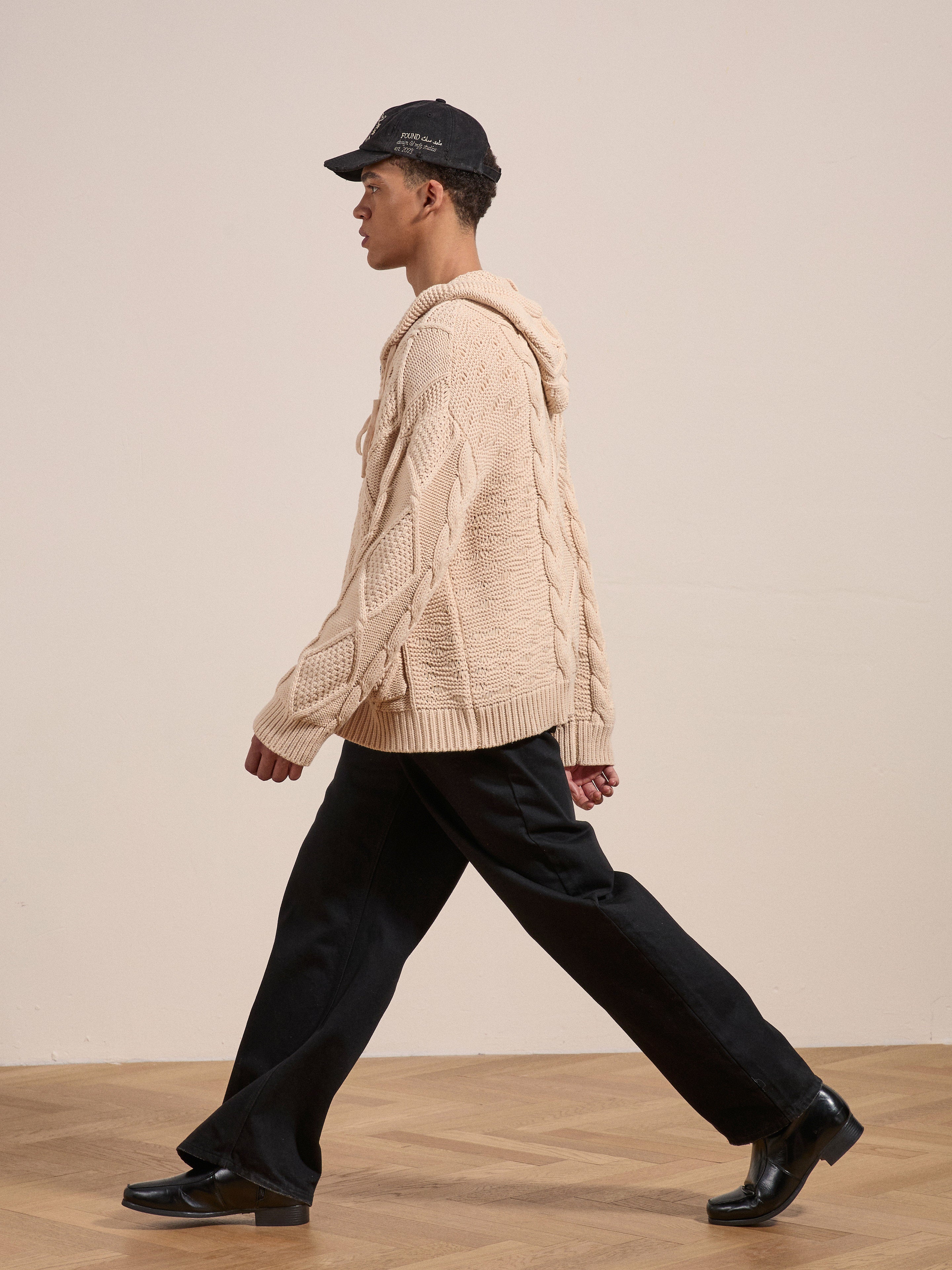 A person in a beige Zip Up Cable Knit Hoodie by Found, paired with black pants and black shoes, is walking sideways against a plain background.