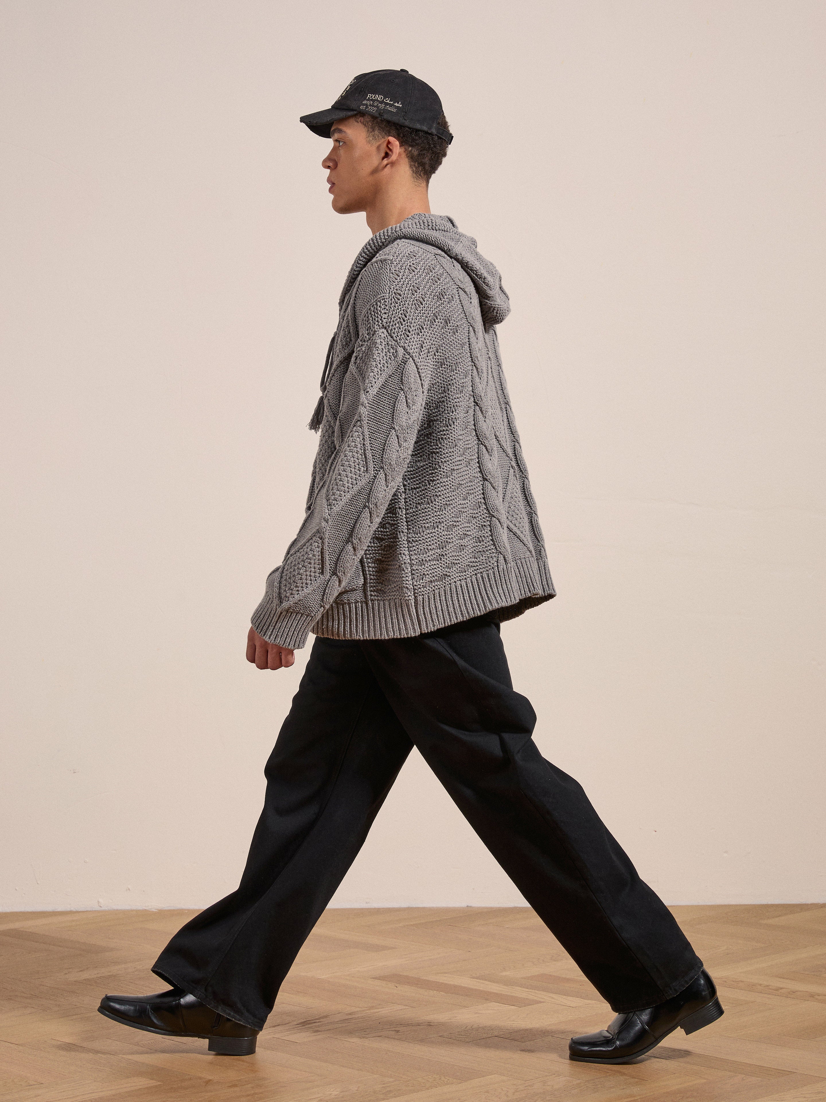 A person dressed in a gray Found Zip Up Cable Knit Hoodie, black pants, and black shoes is walking against a plain background, exuding a cozy look and feel.