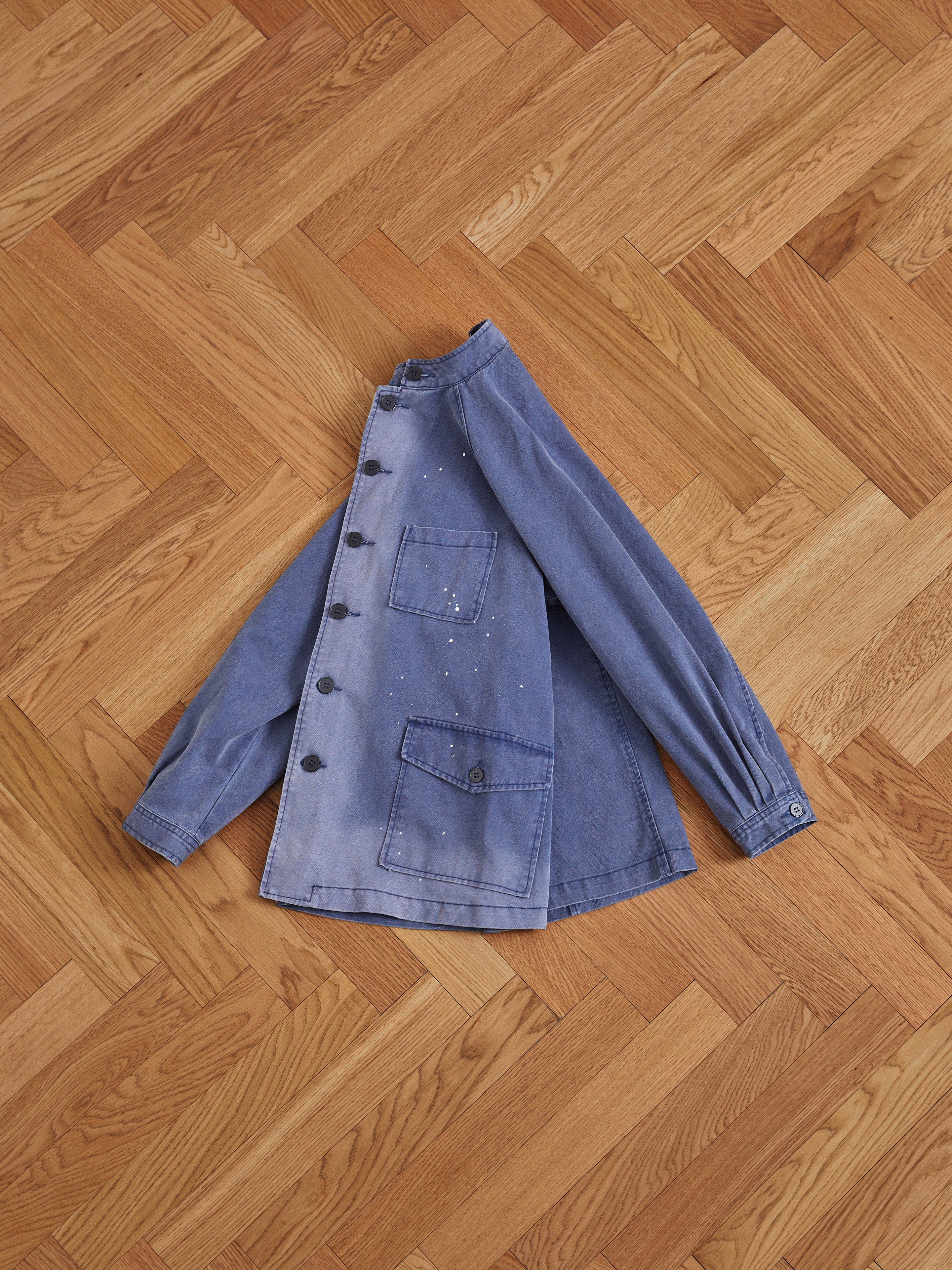 A Found River Painters Chore Coat denim jacket in a classic French silhouette rests on a wooden floor, featuring pleated sleeve cuffs.