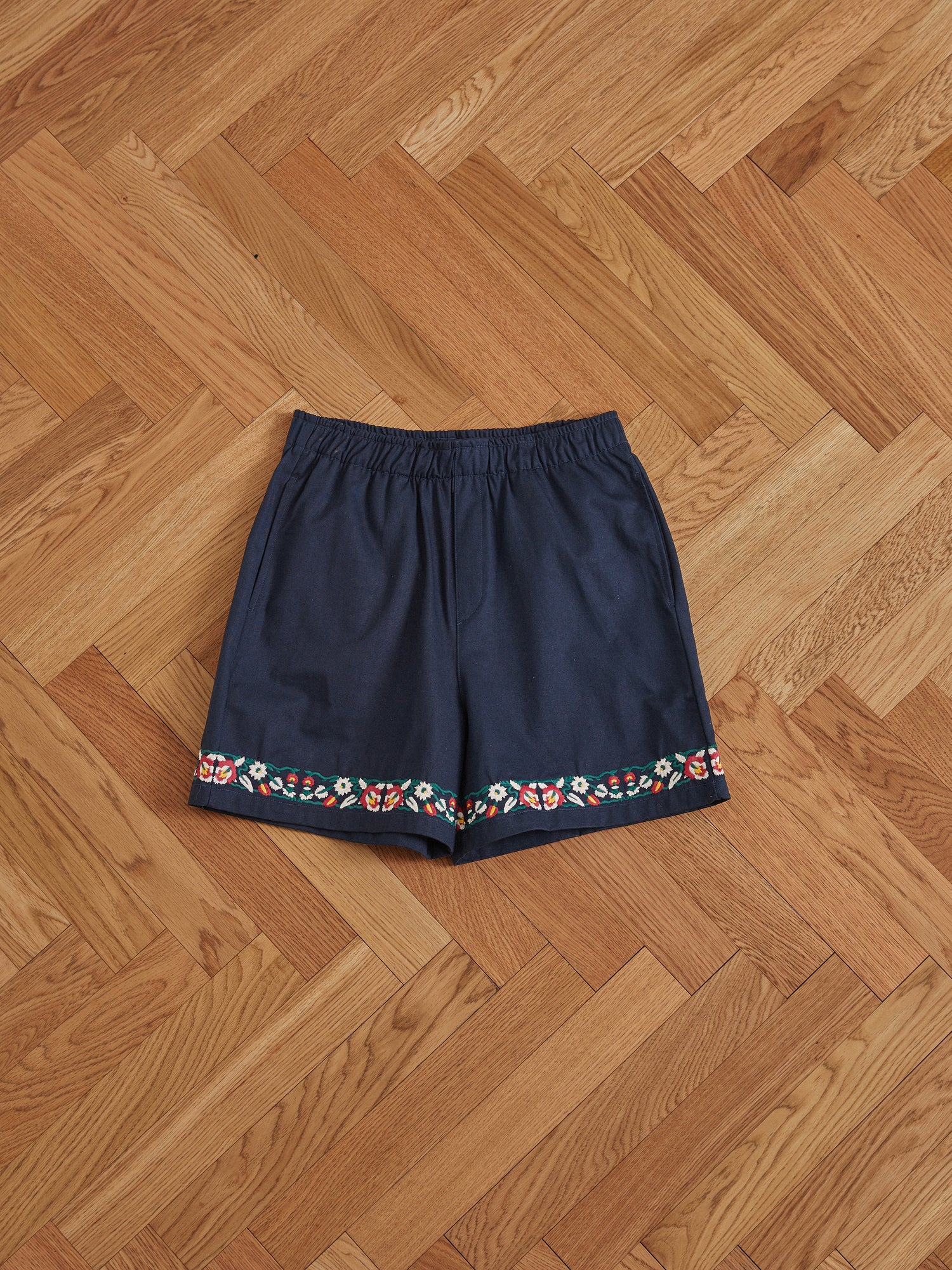 Found Navy blue Horse Equine Twill Shorts with colorful floral embroidery along the hem, lying flat on a herringbone patterned wooden floor.