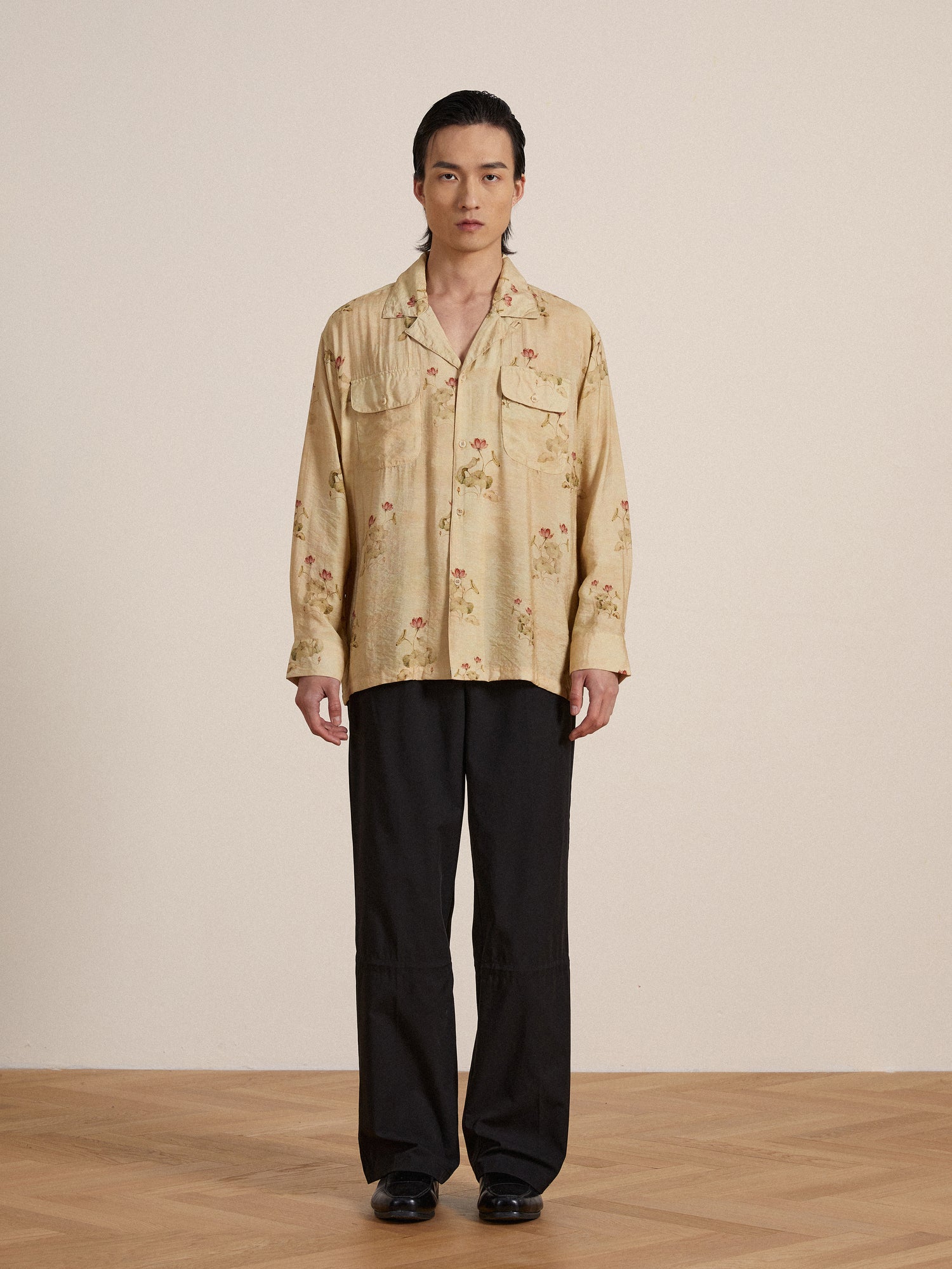 Asian man in a studio wearing a beige floral shirt and black Found Tencel pleated pants, standing with a neutral expression.