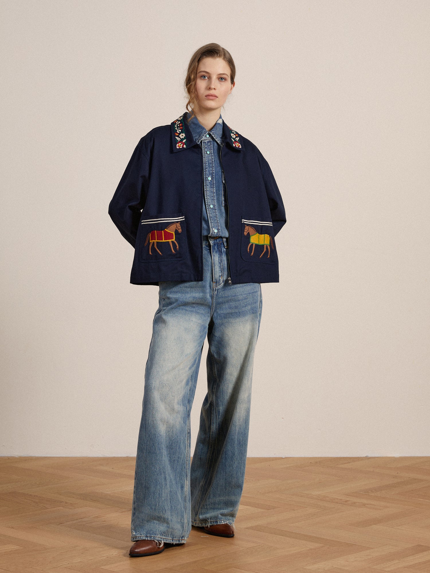 A woman wearing a denim shirt under a Found Horse Equine Work Jacket with detailed embroidery and wide-leg jeans stands in a neutral-toned room.
