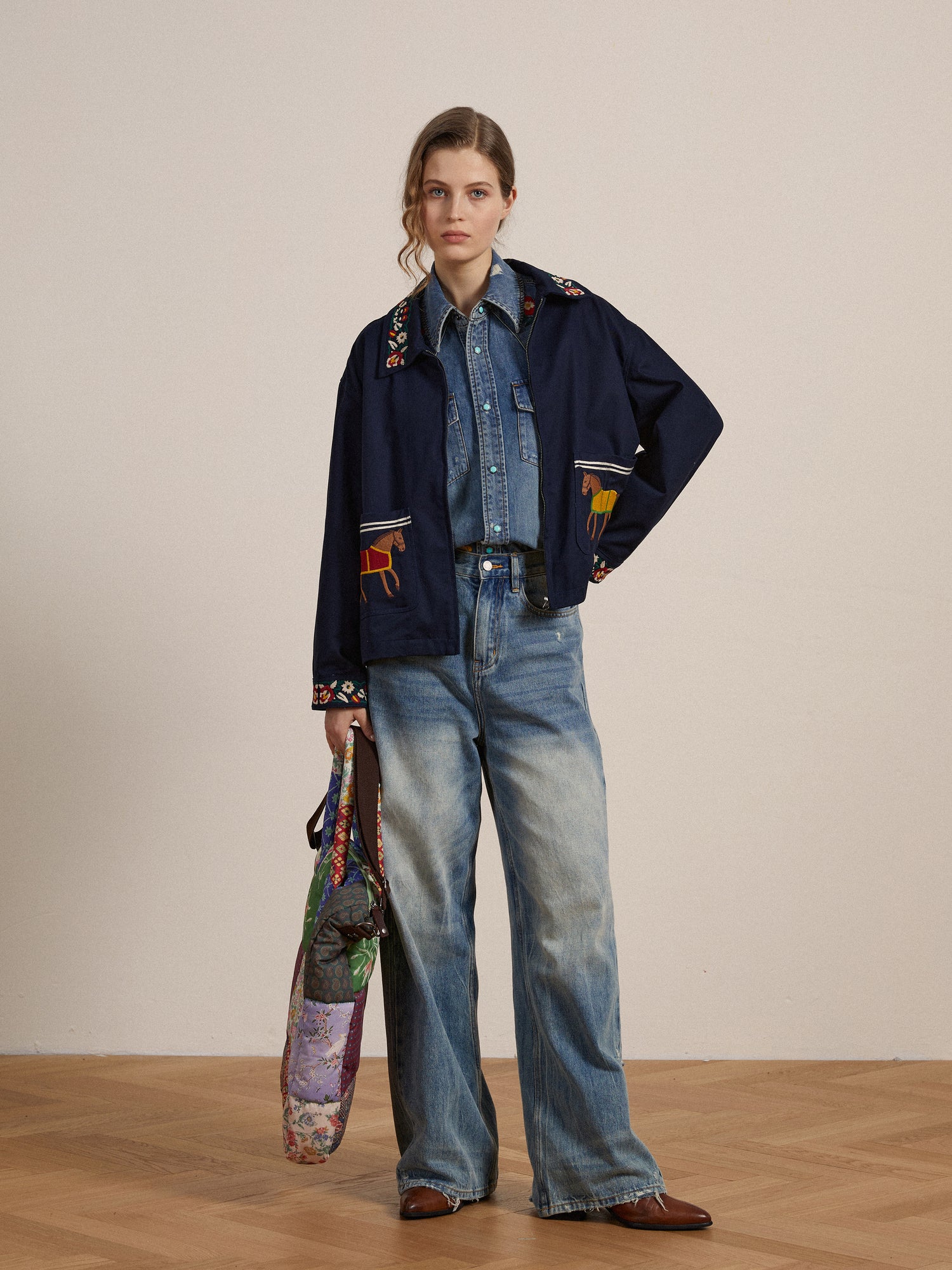 A woman in a denim outfit stands with her hands in her pockets, holding a colorful bag. She wears wide-legged jeans adorned with detailed embroidery, a Found Horse Equine Work Jacket, and brown shoes.