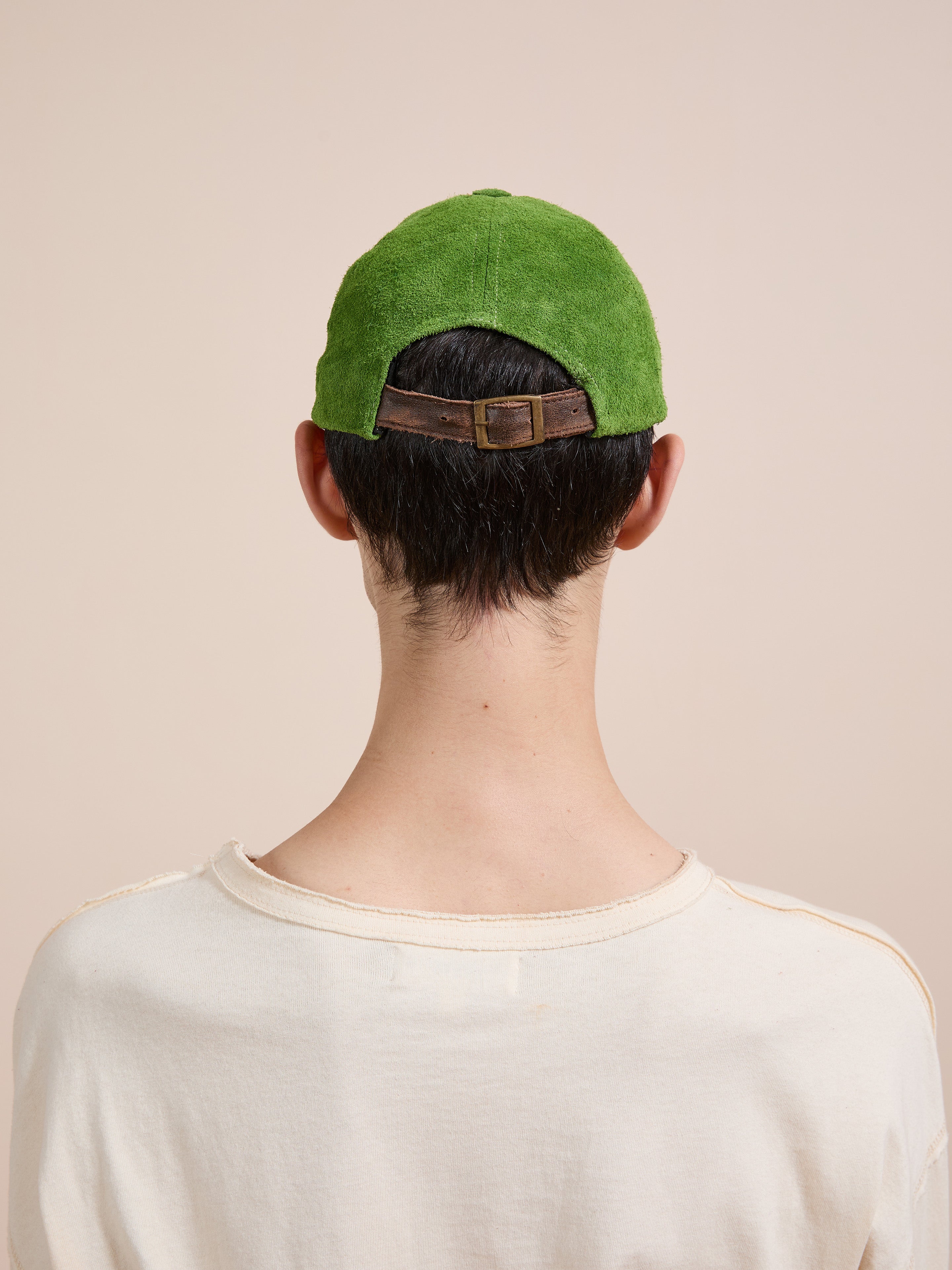A person, seen from behind in a cream shirt, wears the Found Suede Leather Contrast Distressed Cap featuring green suede, a distressed brown leather brim, and an adjustable leather strap.