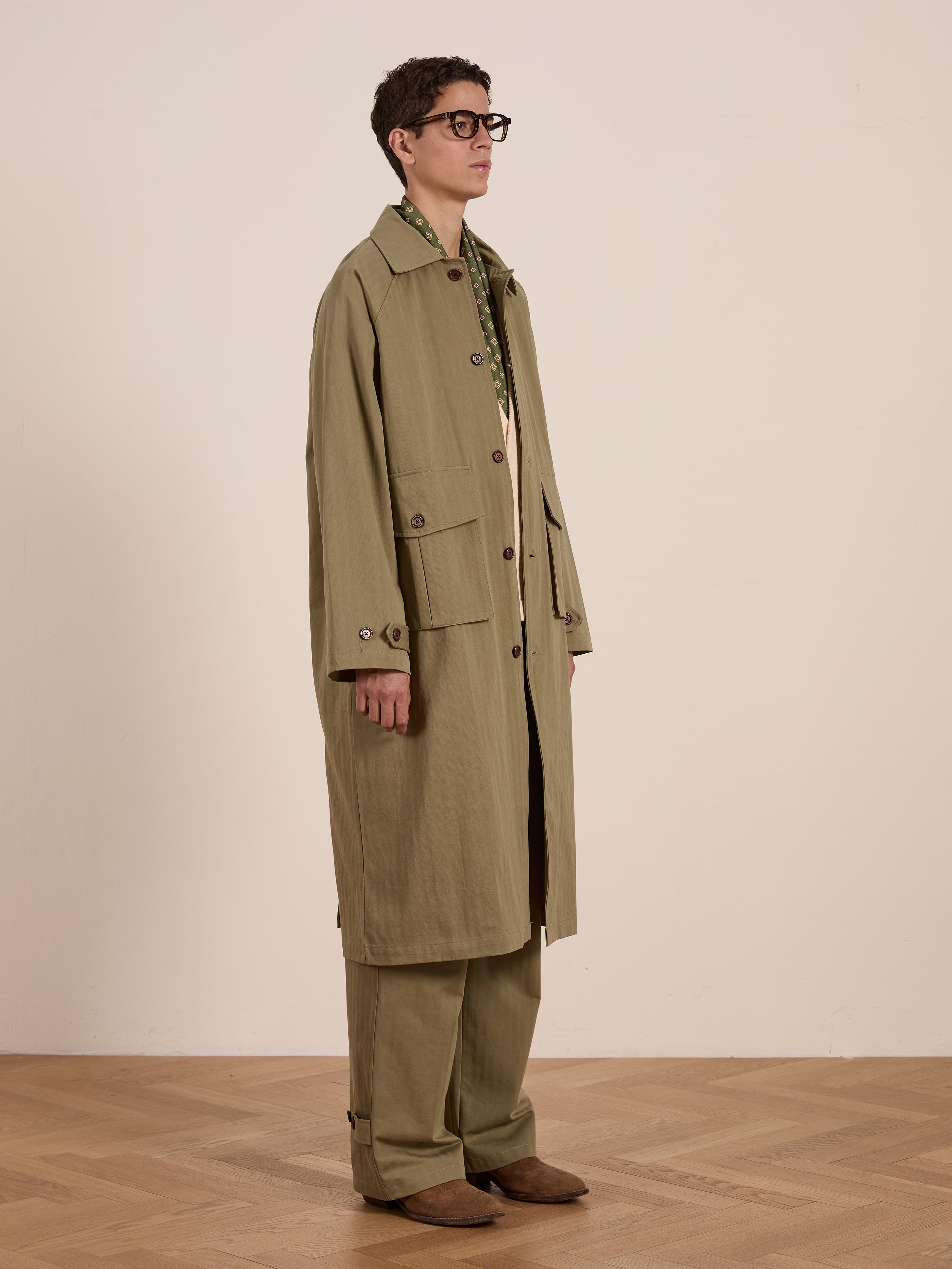 A person is standing sideways on a wooden floor wearing an olive green Herringbone Twill Trench Coat by FOUND, paired with matching pants and a patterned shirt.