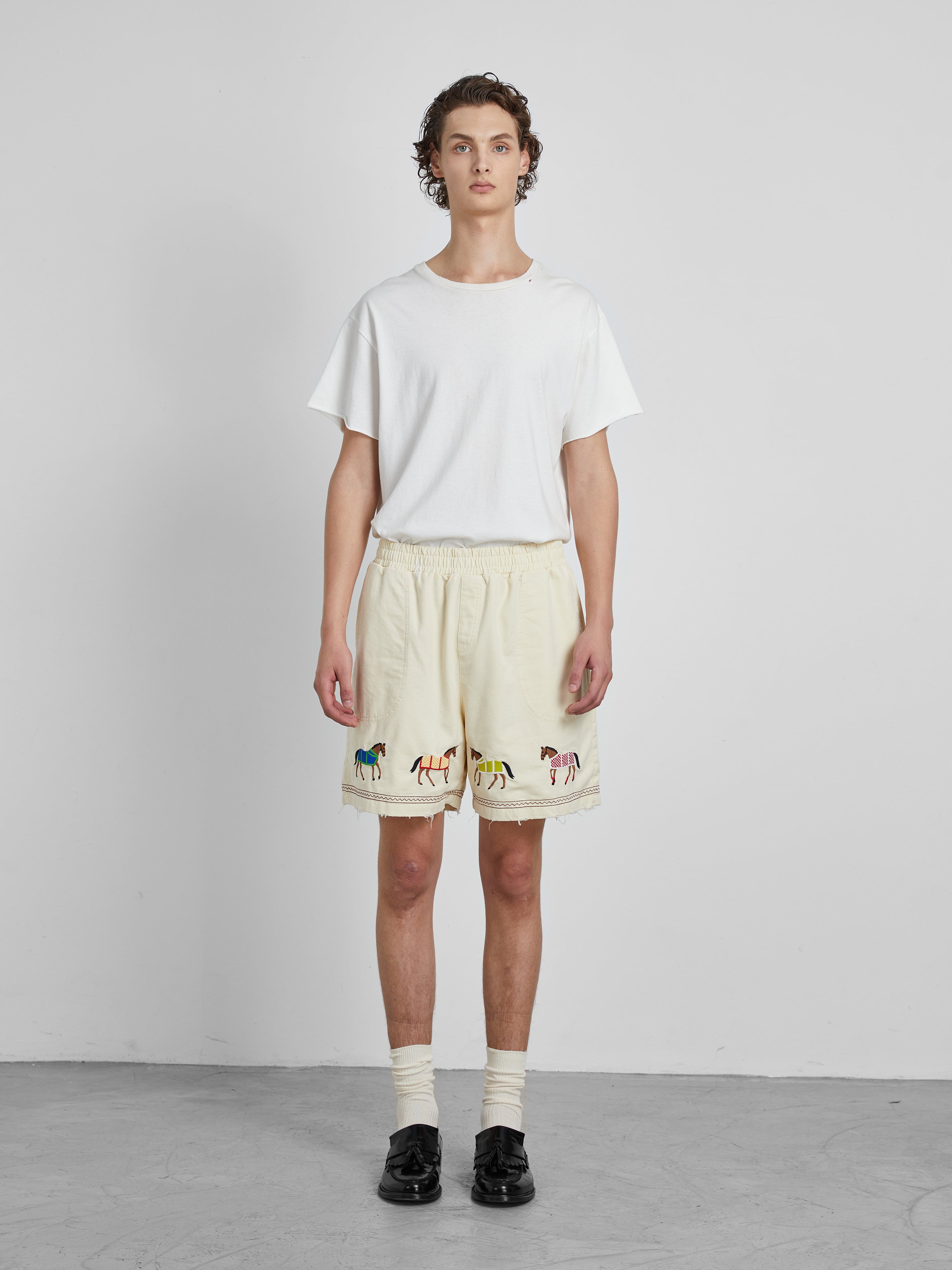 A person stands against a plain white background wearing a white T-shirt, Profound's Embroidered Horse Shorts in cream, cream socks, and black loafers.