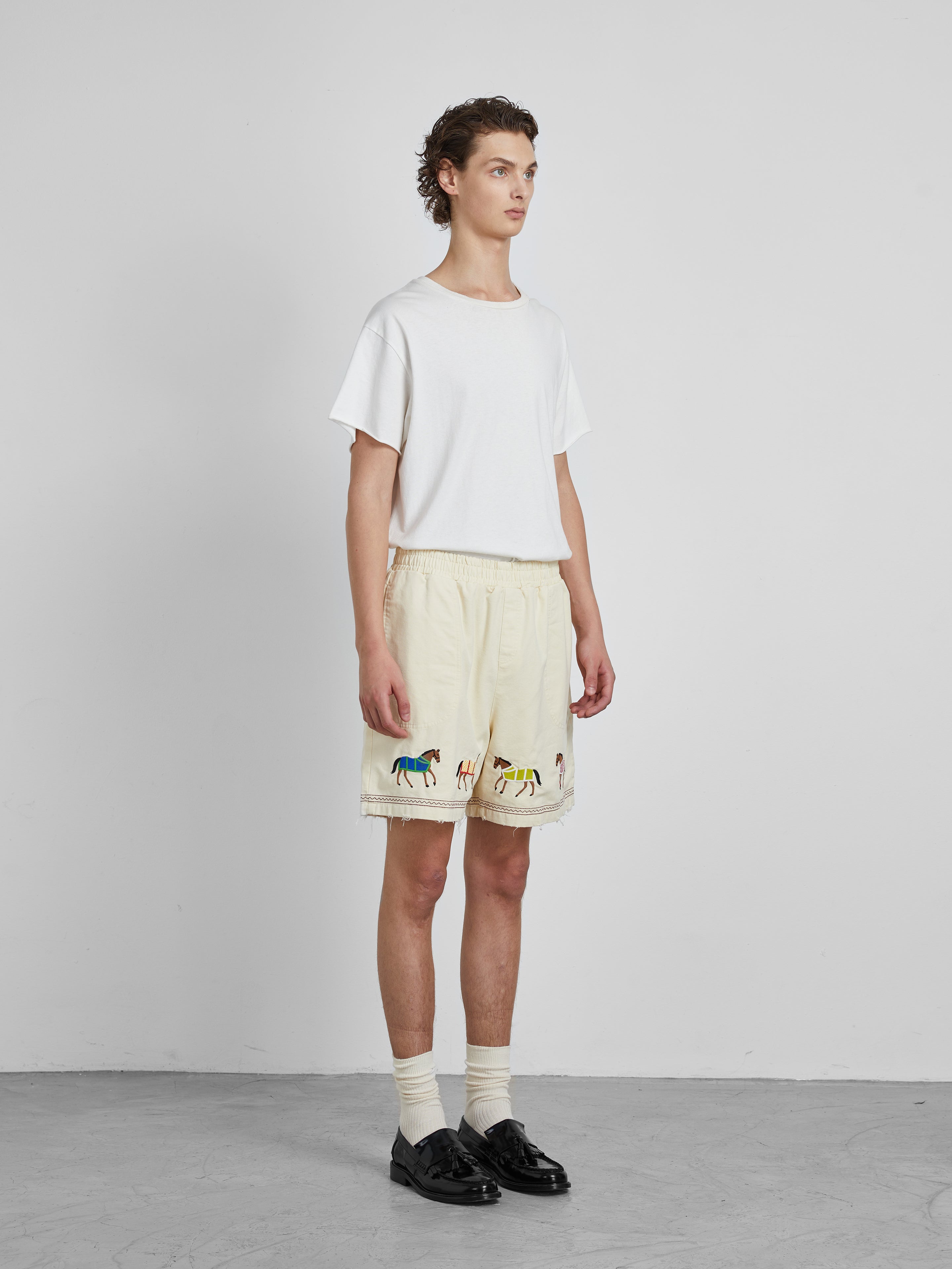 A person in a white t-shirt and Profound's Embroidered Horse Shorts stands against a plain white wall, wearing socks and black shoes.