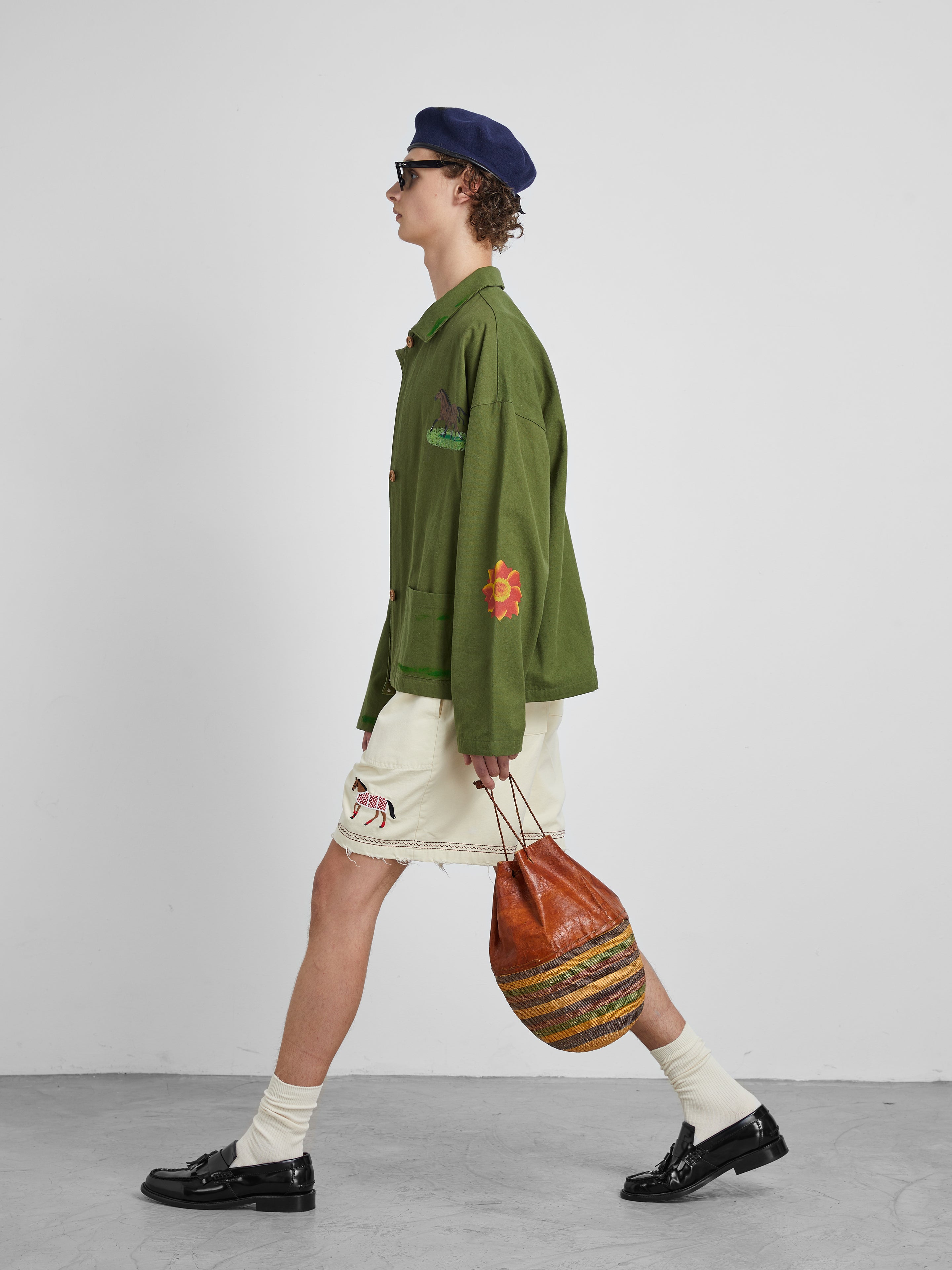 Someone wearing a Profound green jacket and the brand's Embroidered Horse Shorts features cotton twill fabric with intricate horse motifs. They carry a striped bag, wear a beret and sunglasses, and complete their look with black shoes against a plain background.