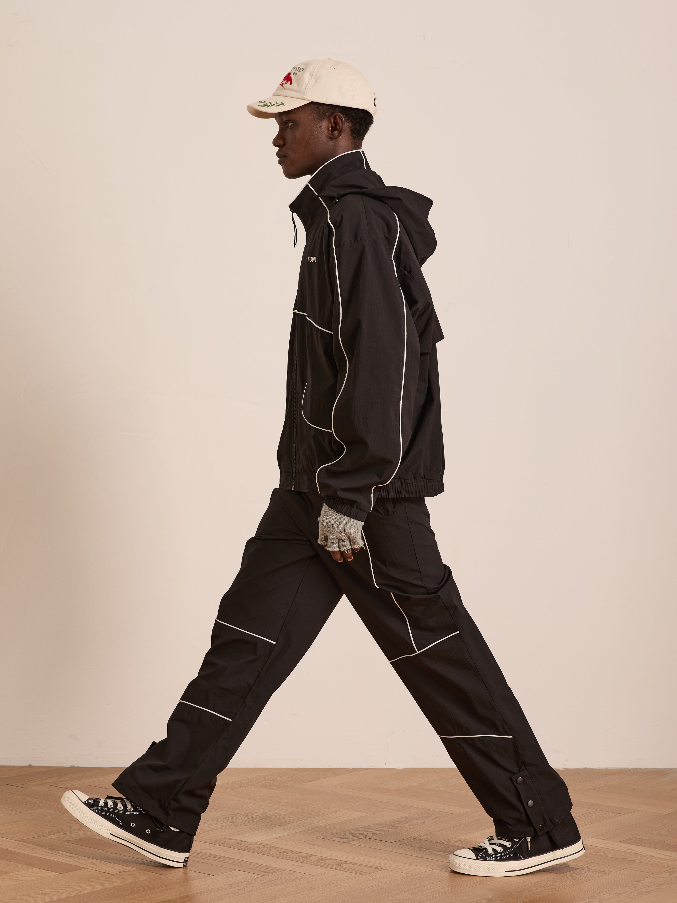 A person walks indoors in a black jacket, white cap, gray gloves, and black Converse sneakers. Their look features Found's Arena Stripe Track Pants—unisex sportswear—standing out against a simple, neutral background.