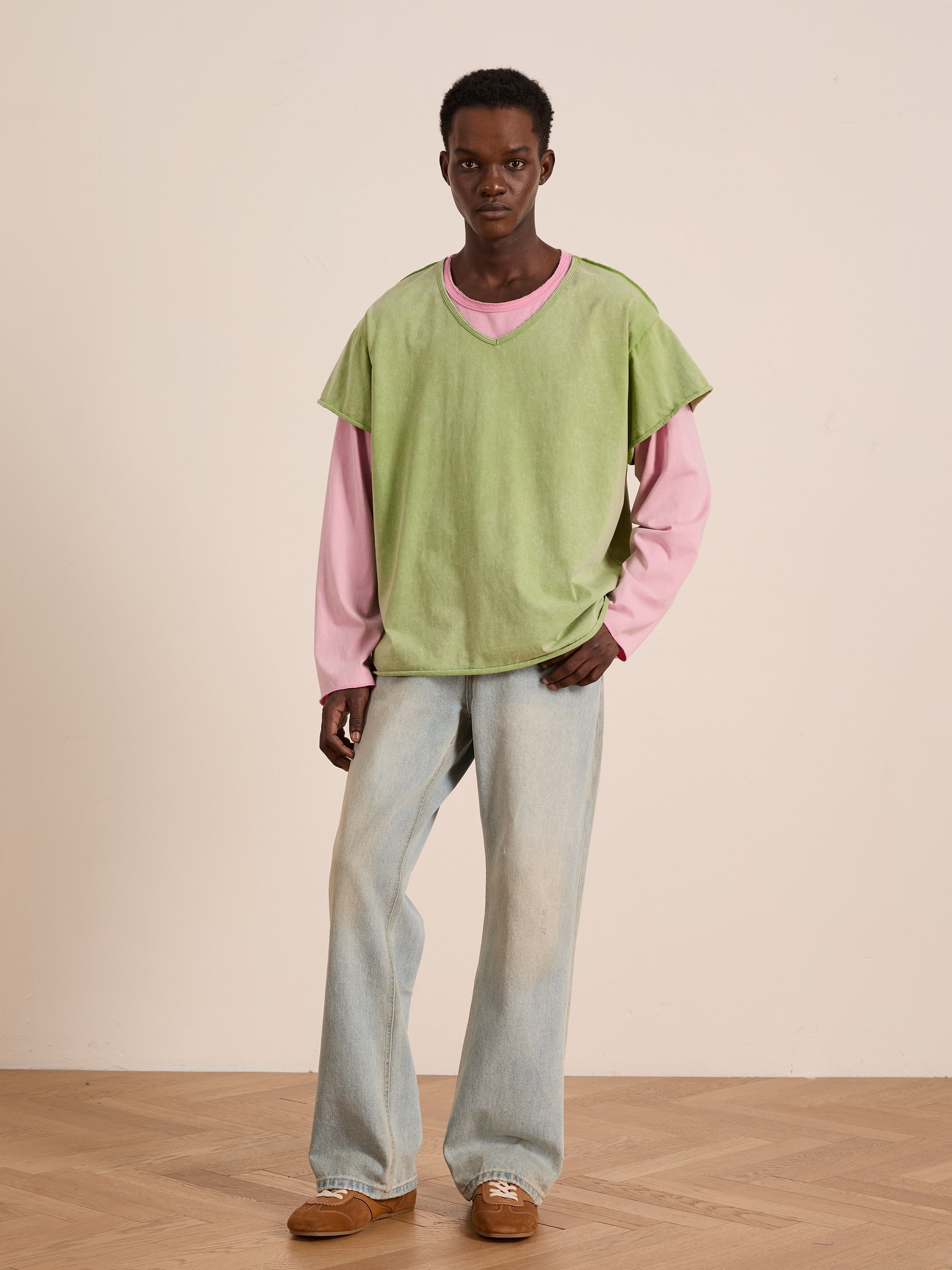 Indoors on a wooden floor, a person wears FOUND's Double Layer LS Tee in light green over a pink unisex long sleeve tee, paired with light blue jeans and brown shoes.