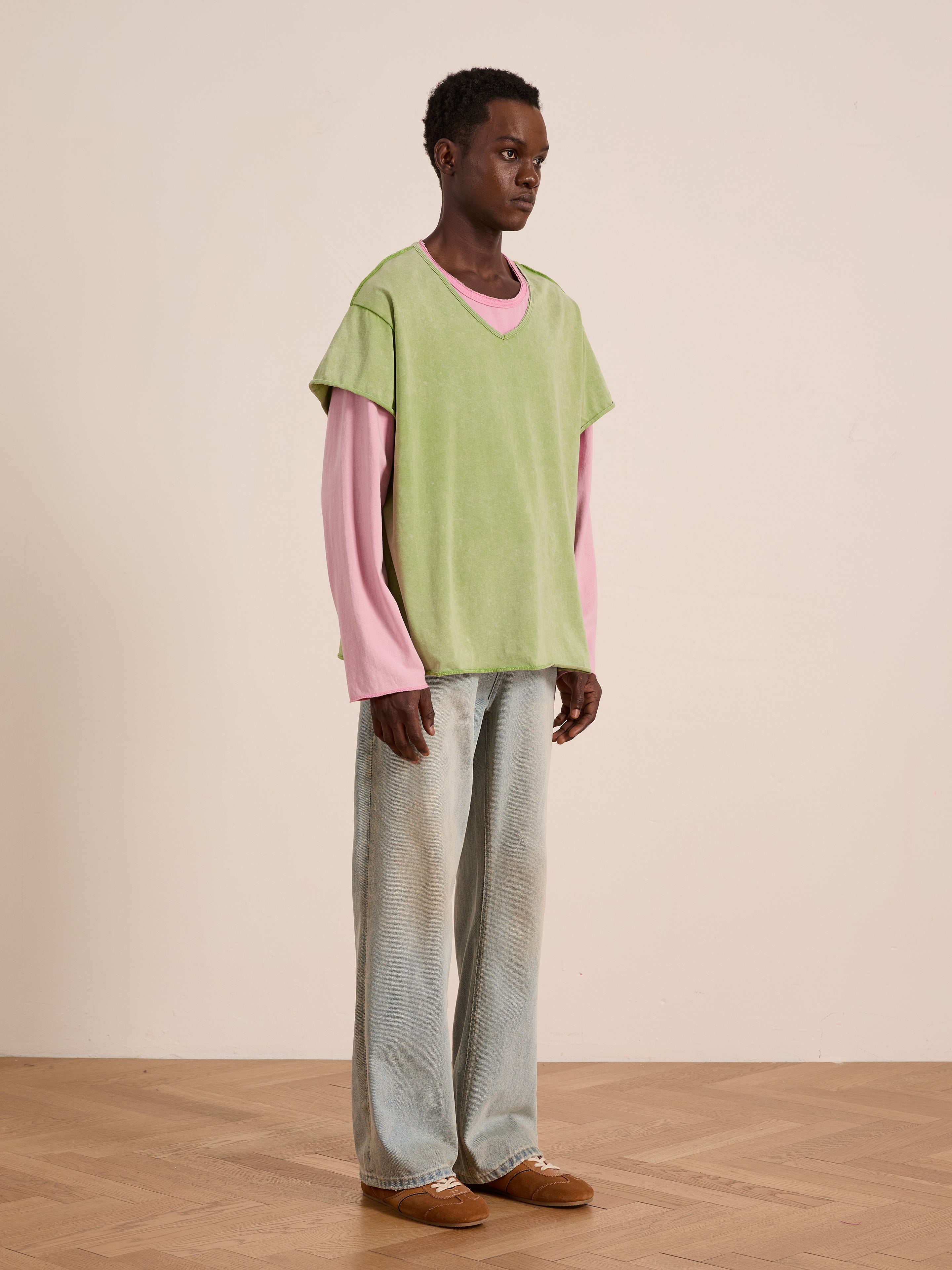 A person stands indoors on a wooden floor against a beige wall, wearing the FOUND Double Layer LS Tee in light green over a pink long-sleeve shirt, with light blue jeans and brown shoes.
