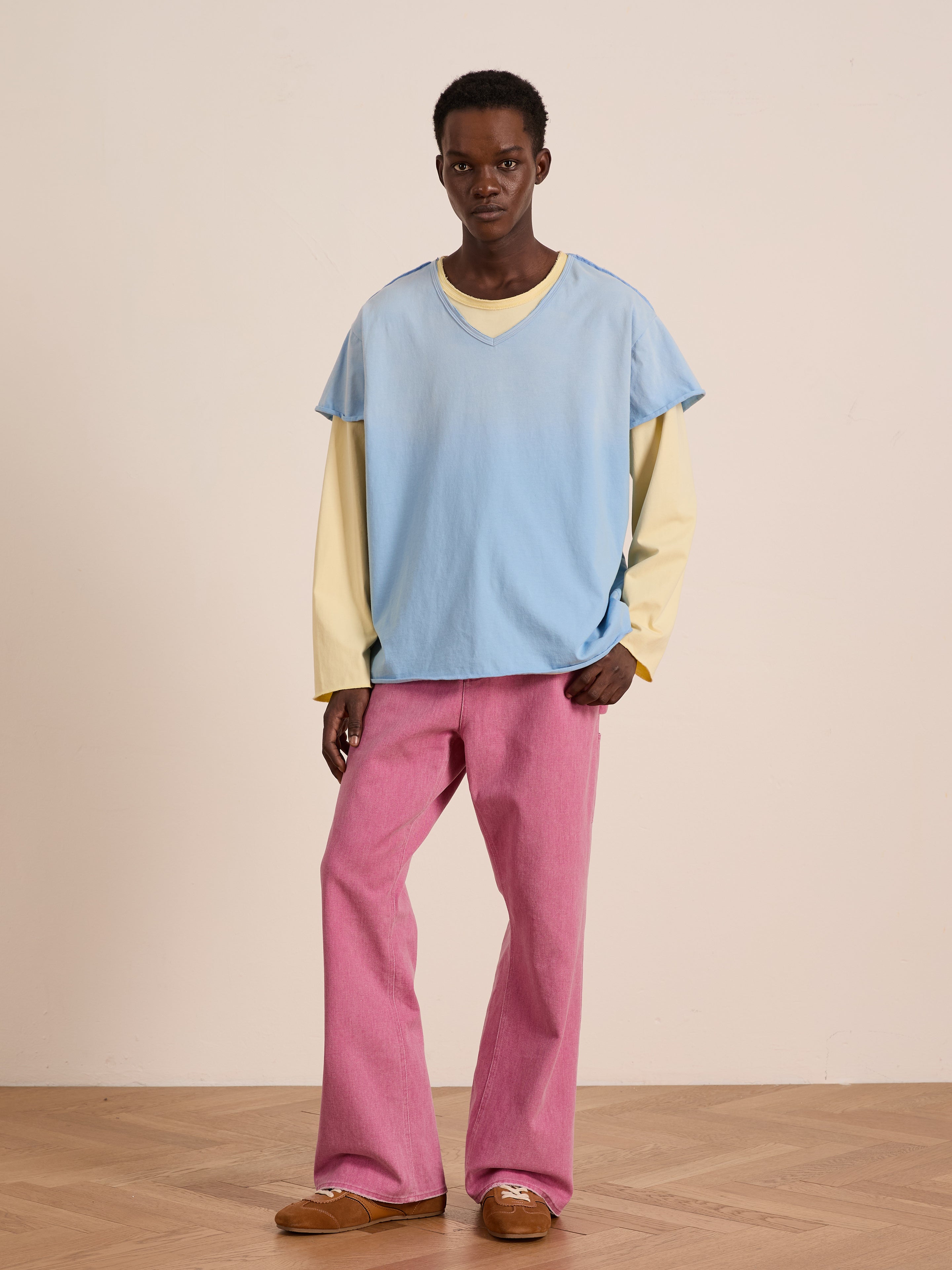 A person wears the FOUND Double Layer LS Tee—light blue short sleeve over yellow long sleeve—matched with pink pants and brown shoes, standing indoors against a beige wall and wooden floor.